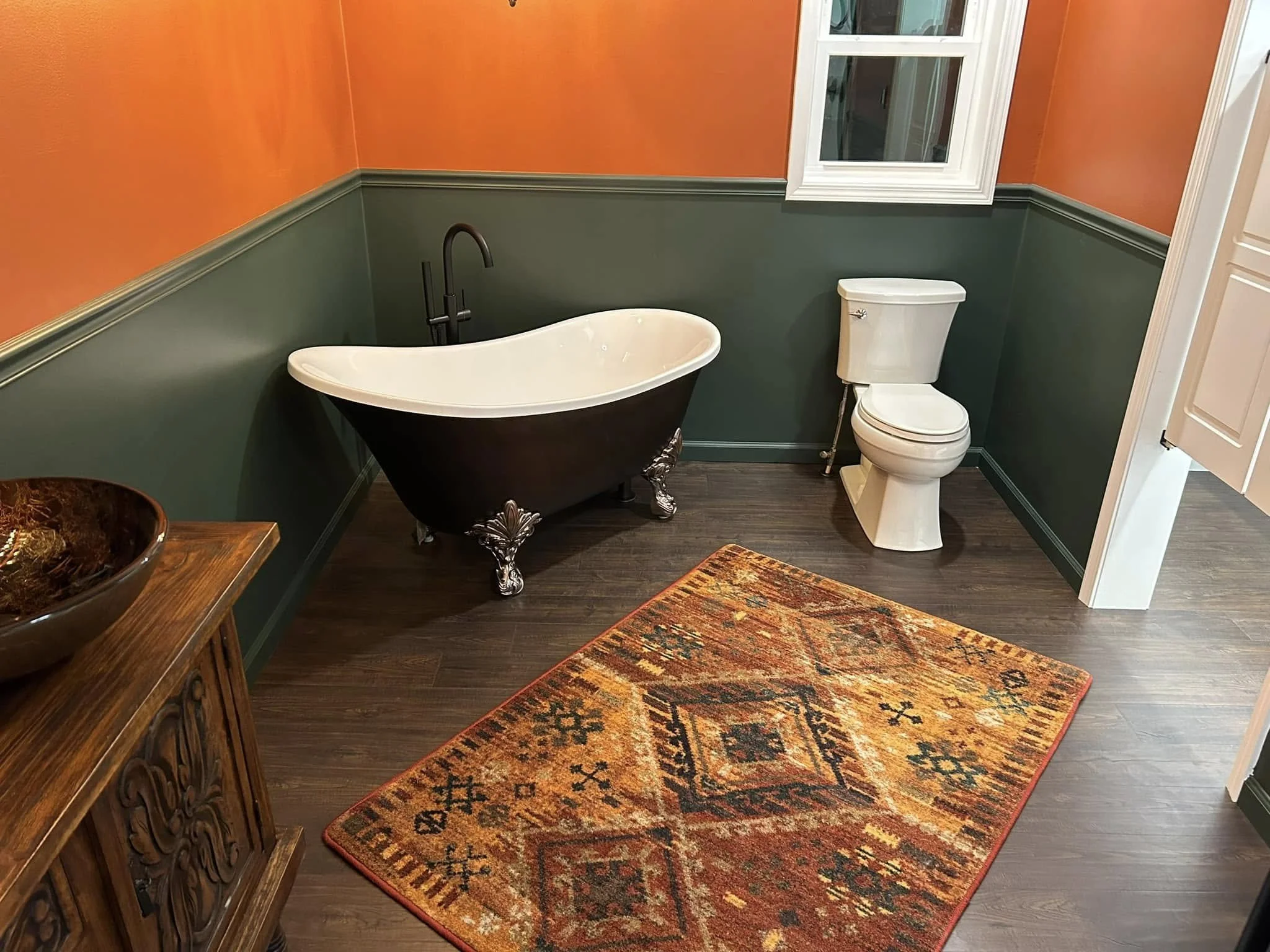 A bathroom with orange and dark green walls, a clawfoot bathtub, a white toilet, a window, a wooden cabinet, and a colorful rug.