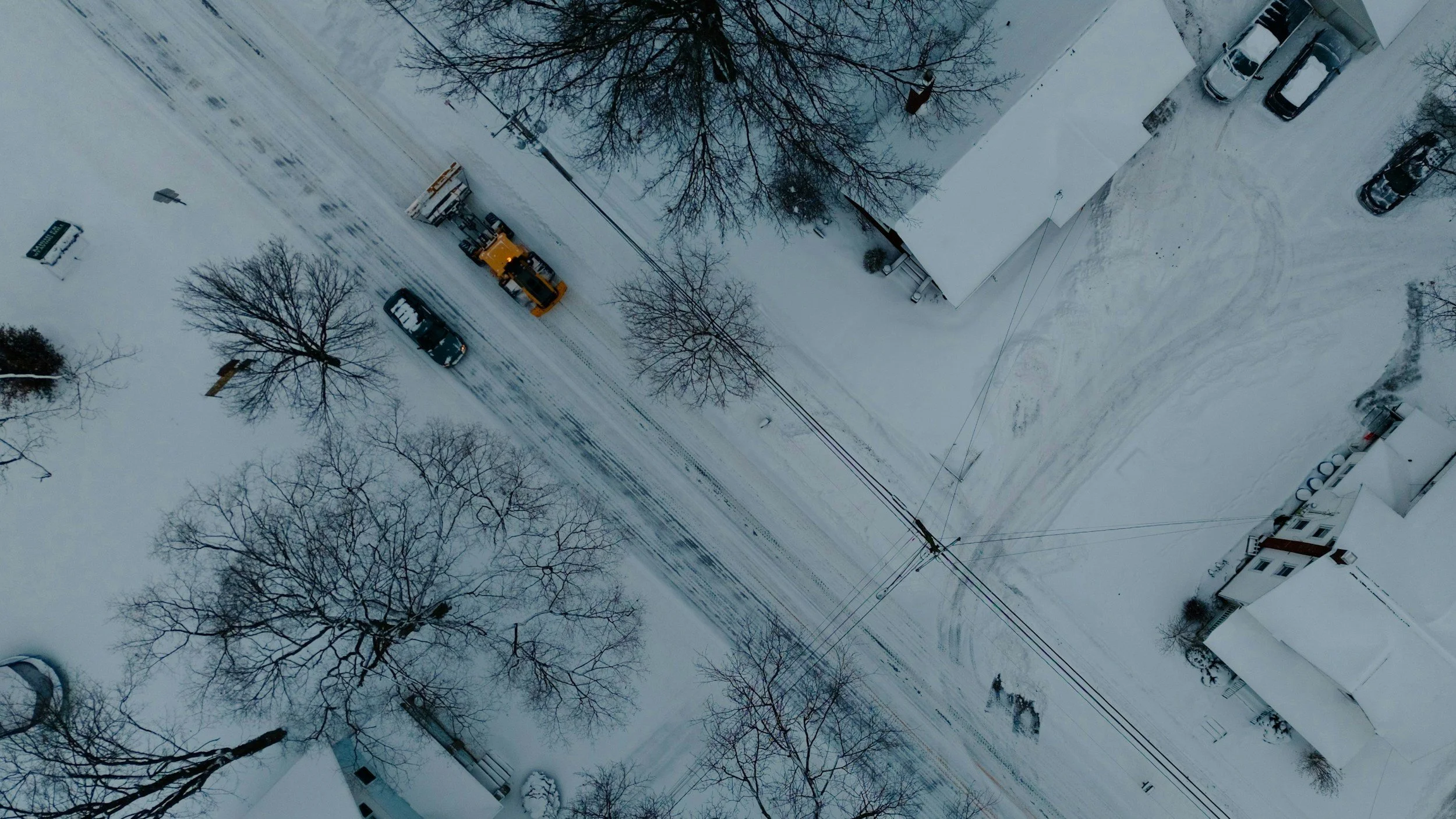 An aerial view of a snowy residential street with parked cars, leafless trees, and a snow-covered house and sidewalk.