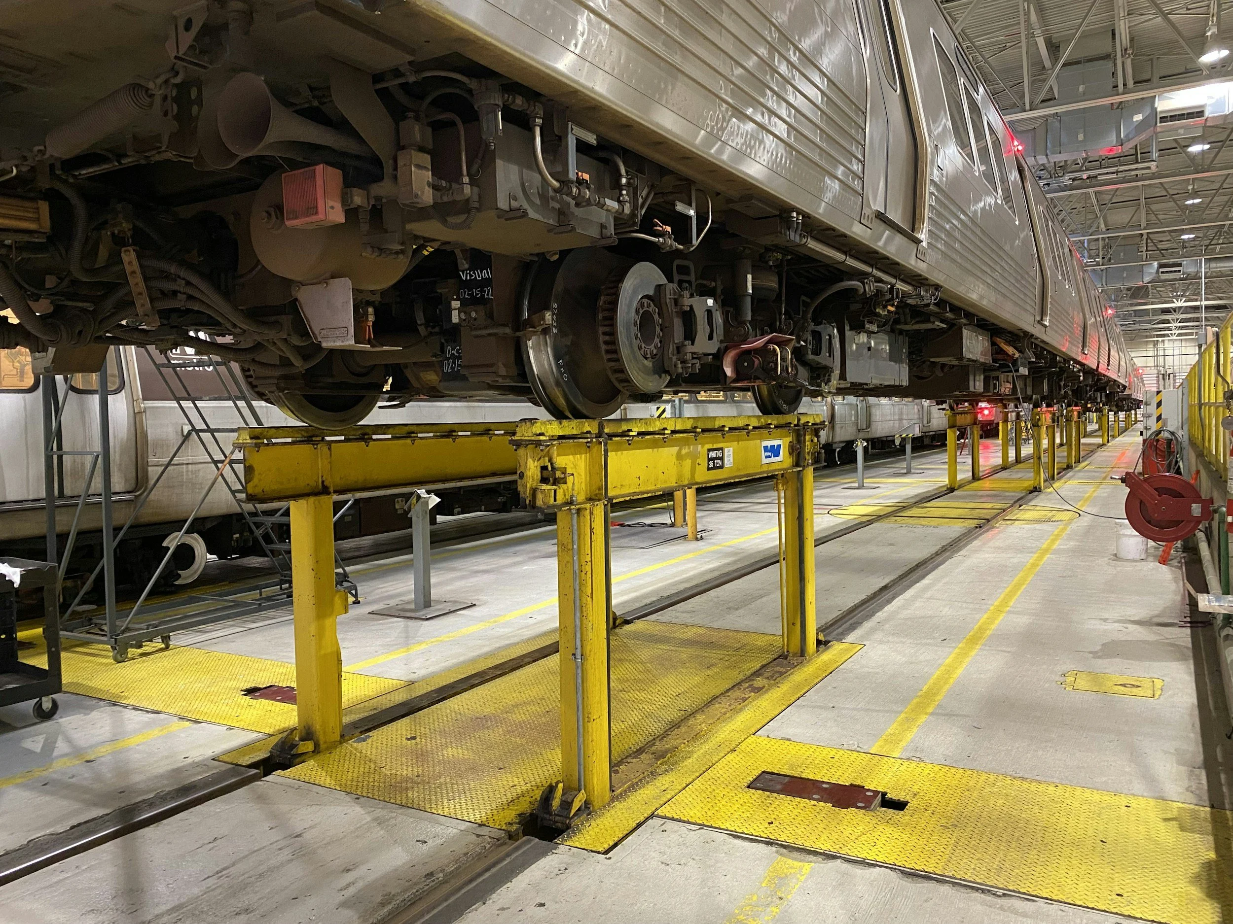 The image shows a train in a maintenance facility lifted on yellow support stands with visible wheels, under a ceiling with exposed metal beams and ductwork. The train is metallic and appears to be undergoing repairs.