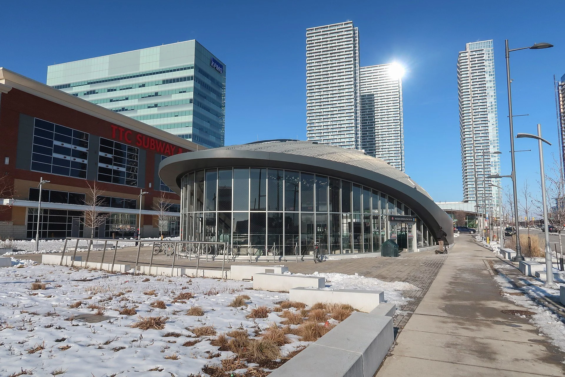 Cityscape featuring modern high-rise buildings with reflective glass windows, a KPMG sign on one building, a TTC Subway entrance with a curved glass roof, leafless trees, street lamps, and snow-covered ground under a clear blue sky.