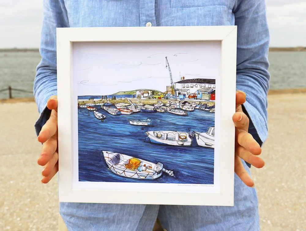 A person holding a framed image of Bullock Harbour, Dalkey with boats, buildings, and water, standing outdoors on Dún Laoghaire Pier.