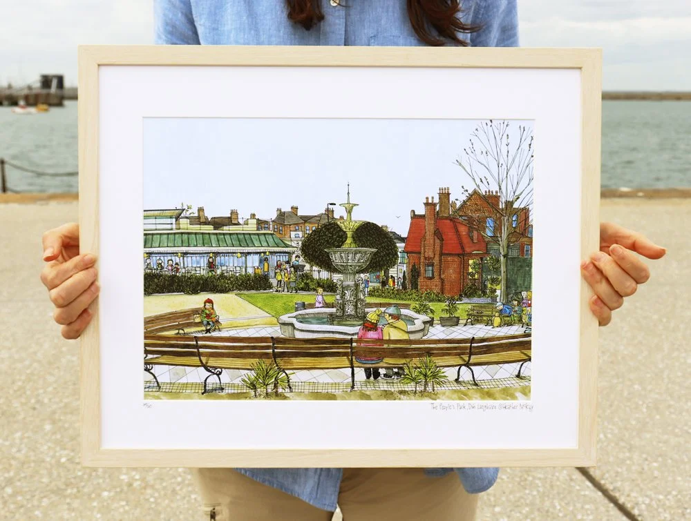 A person holding a framed image of The People's Park, Dún Laoghaire with benches, fountain, trees, and buildings in the background.
