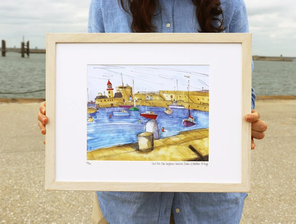 A person holding a framed illustration of Dún Laoghaire Harbour with boats and a stone wall.