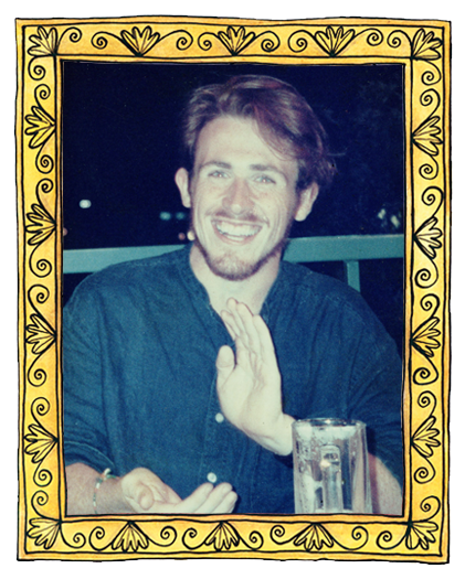 A young man smiling, with long brown hair and a beard, sitting at a table with a glass of beer in front of him, framed by a decorative yellow border with floral designs.