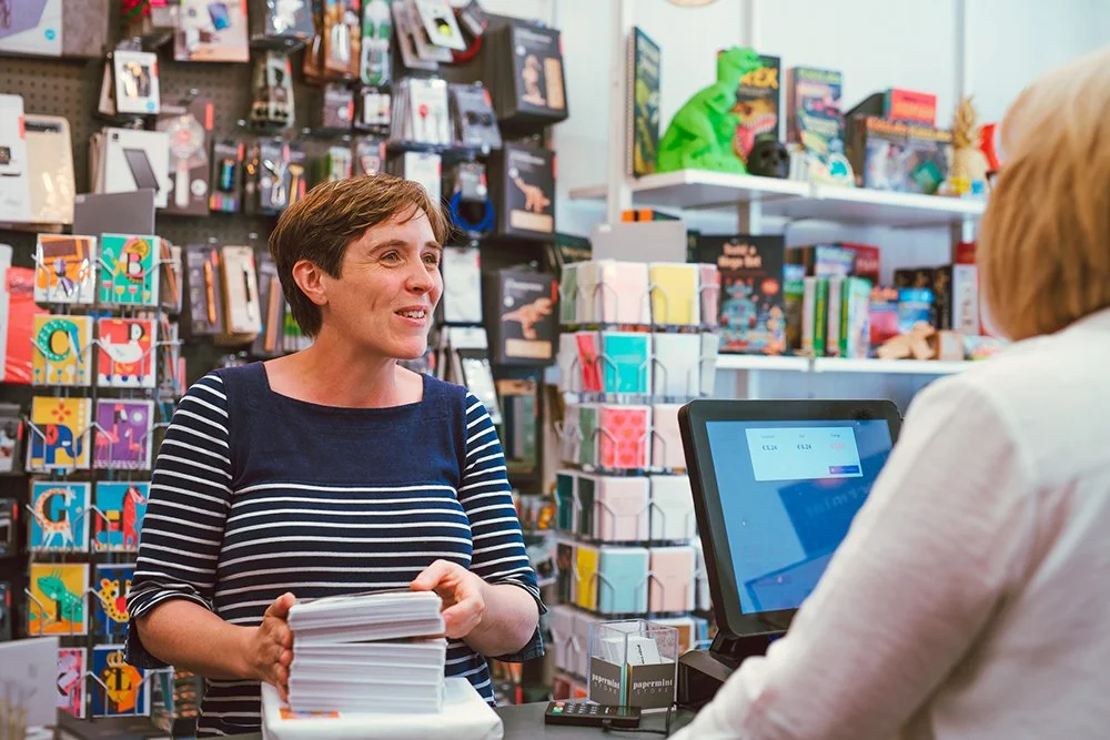 A woman shopping at a bookstore counter with a cashier, surrounded by colorful greeting cards, books, and stationery.