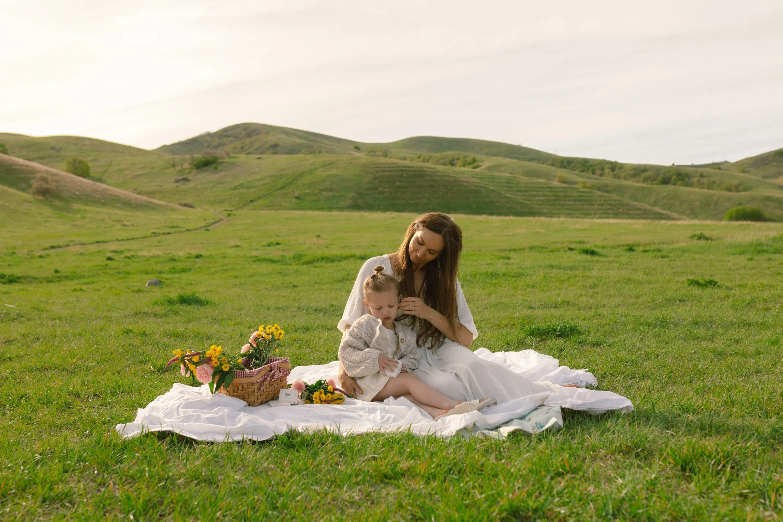 A woman and a young girl sitting on a blanket in a grassy field with rolling hills in the background, surrounded by picnic items and flowers.