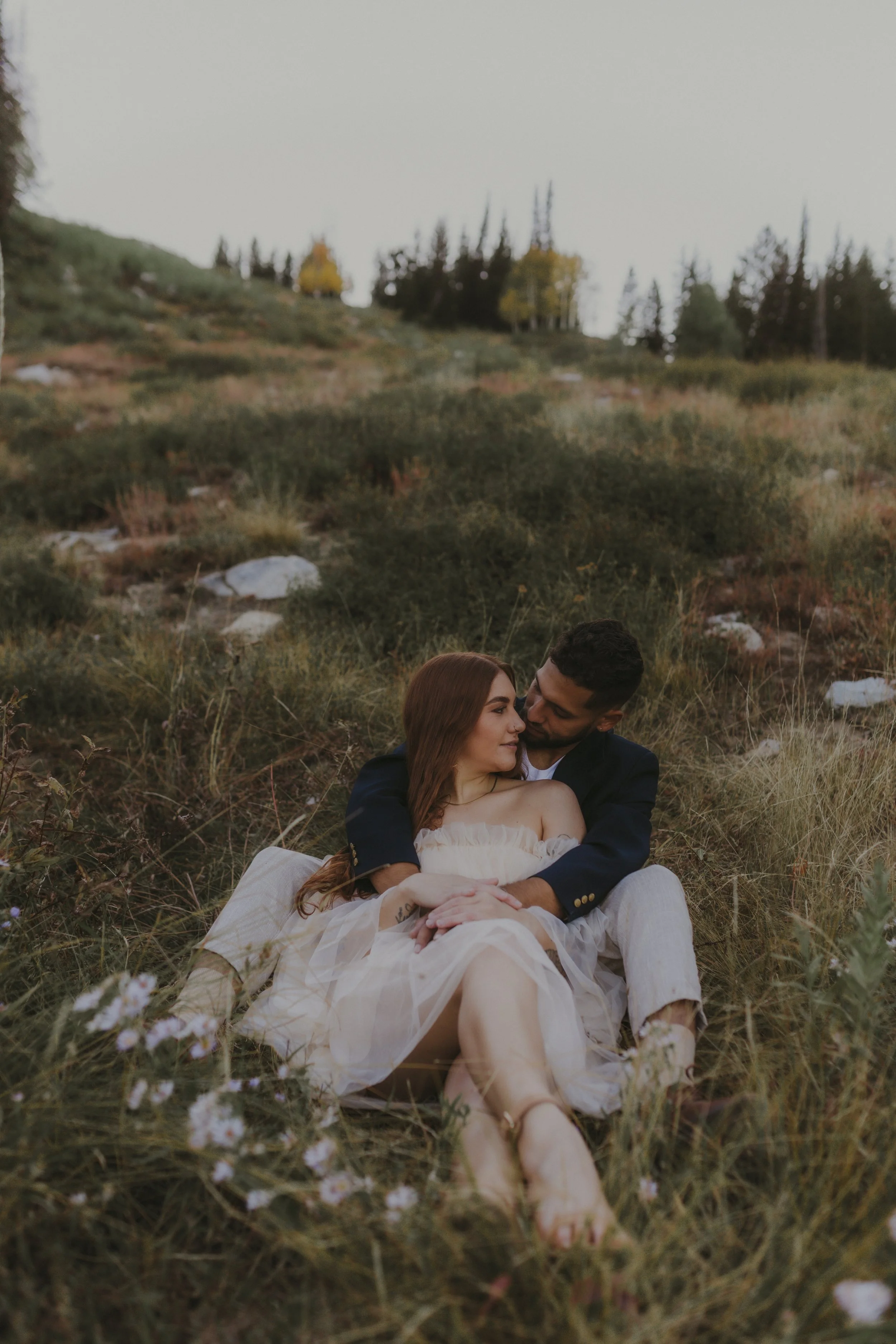 A couple sitting on the grass in a field, embracing each other with a backdrop of trees and rock formations in a natural outdoor setting.