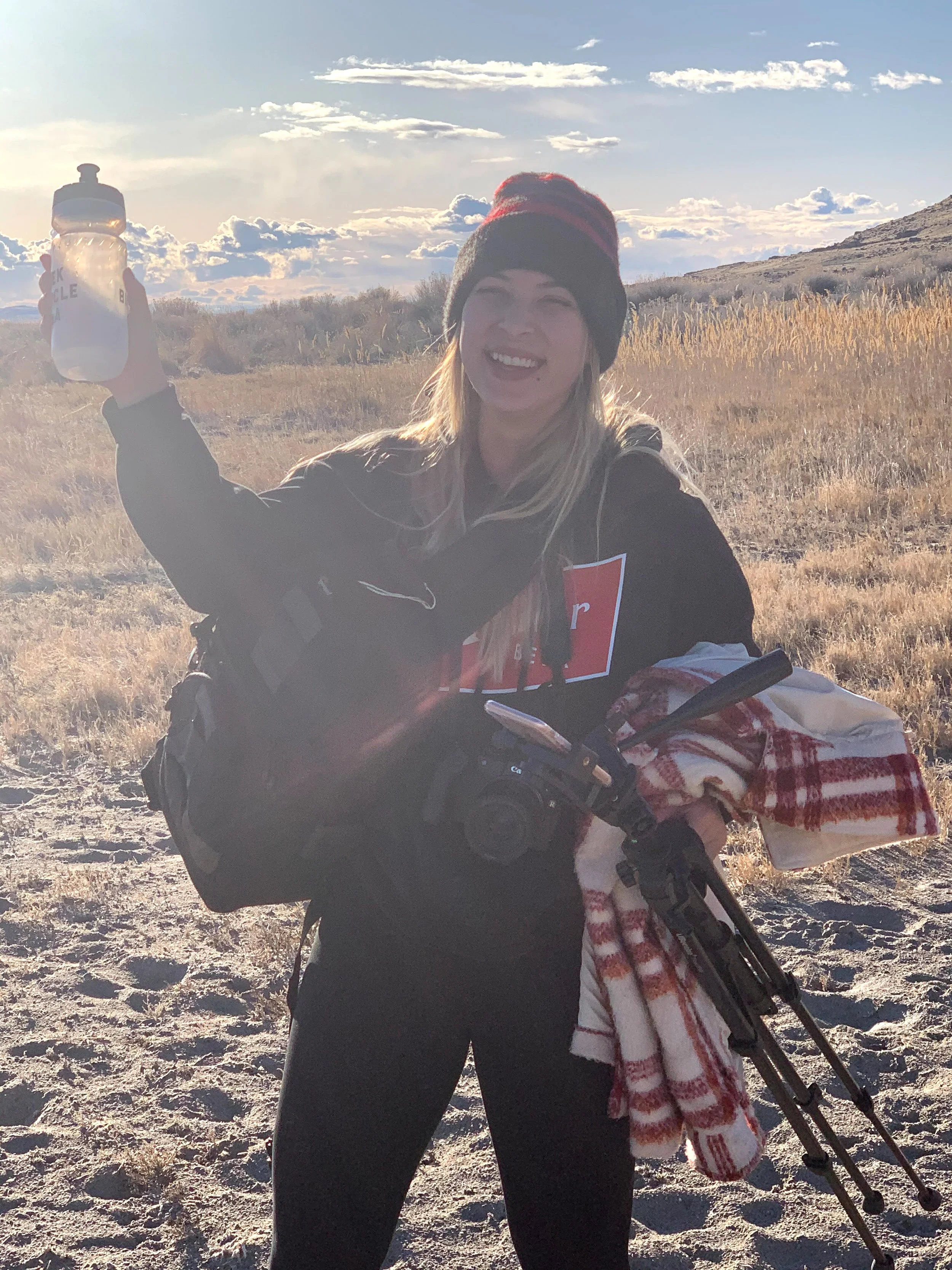 A woman smiling and holding a water bottle outdoors in a desert landscape with mountains, grasses, and clouds in the background.