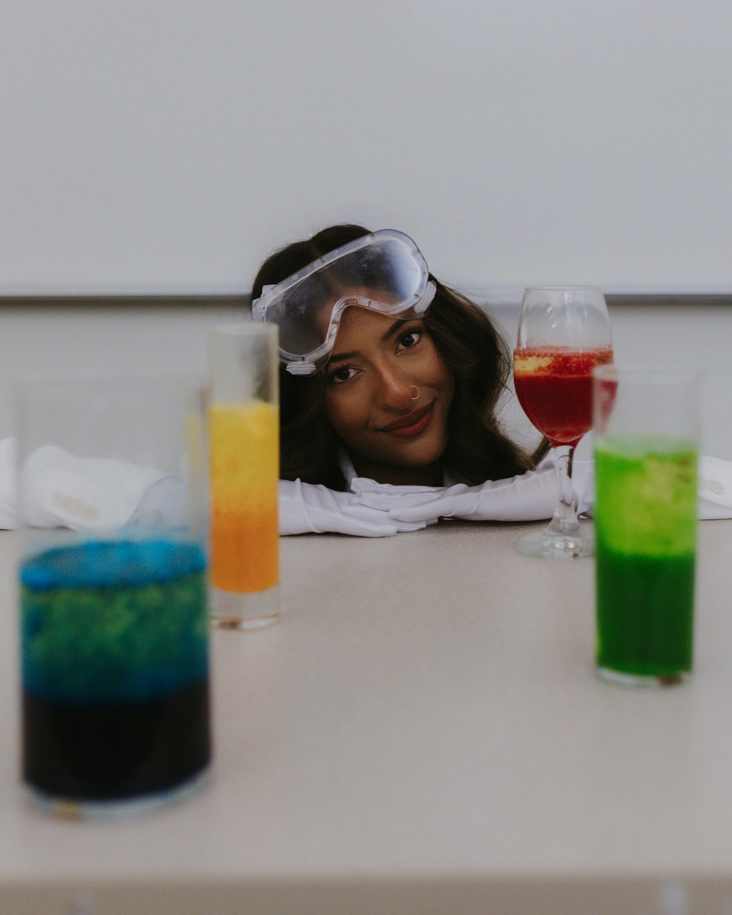 Woman wearing protective goggles, looking at various scientific liquids in test tubes on a table.