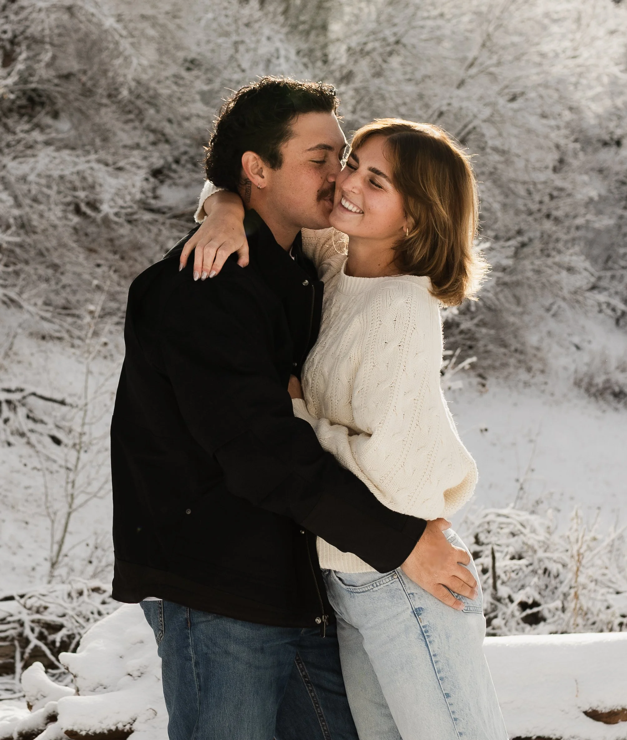 A couple embracing outdoors in a snowy landscape, with snow-covered trees in the background, smiling and enjoying wintertime together.