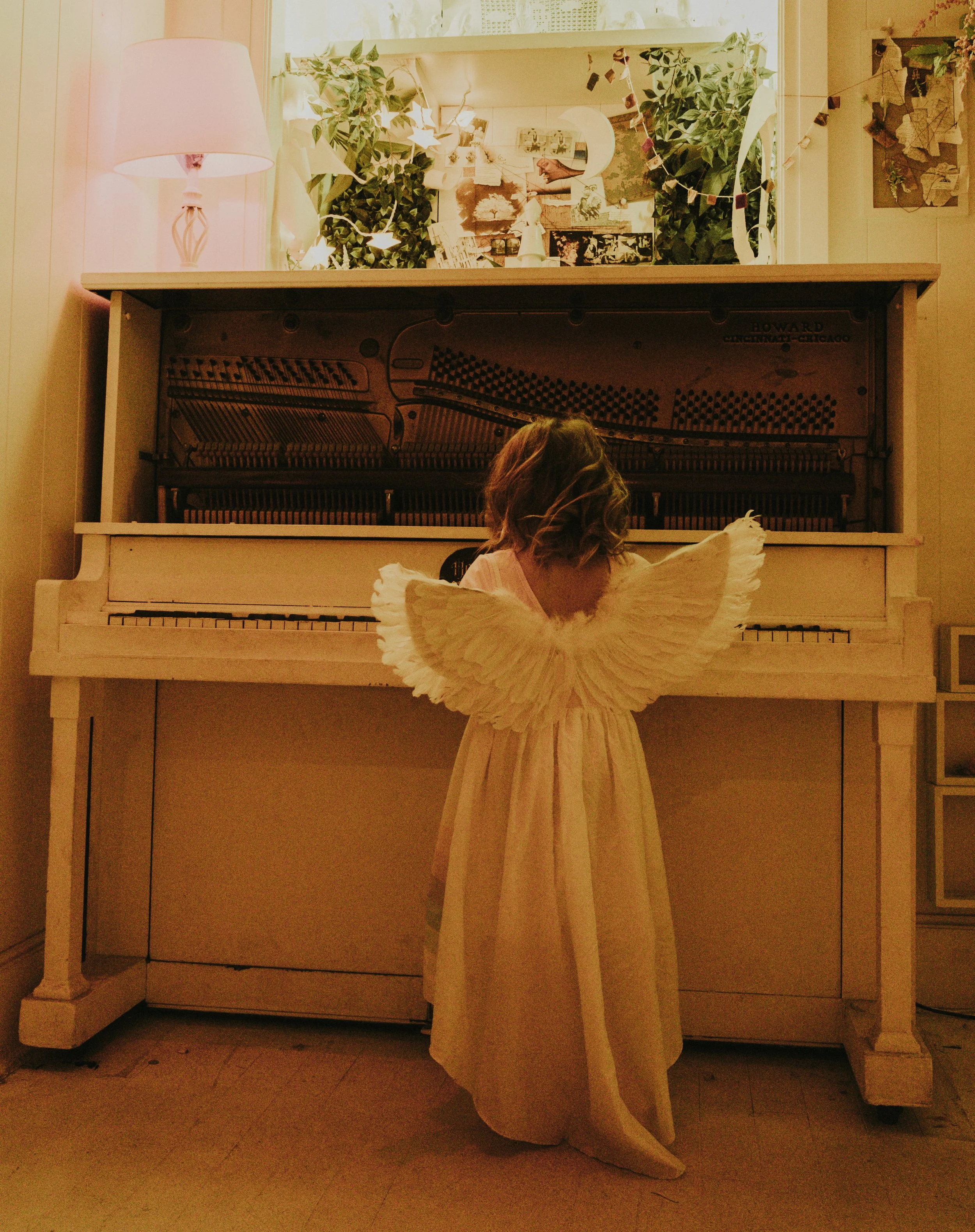 A person dressed as an angel with large white feathered wings and a flowing white gown standing in front of a white upright piano. The person's head is turned away from the camera.