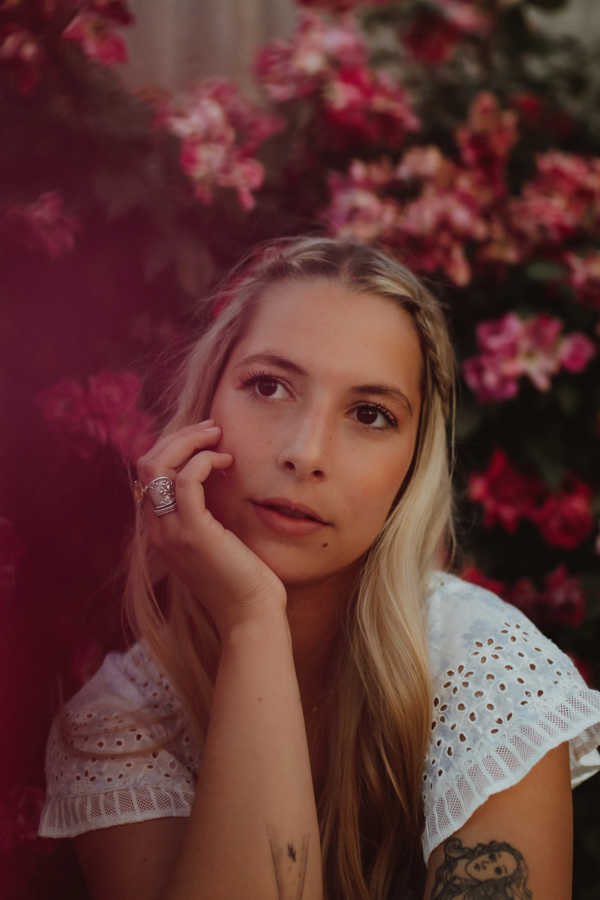 A young woman with long blonde hair and a tattoo of a lady's face on her arm, resting her chin on her hand, with pink flowers in the background.