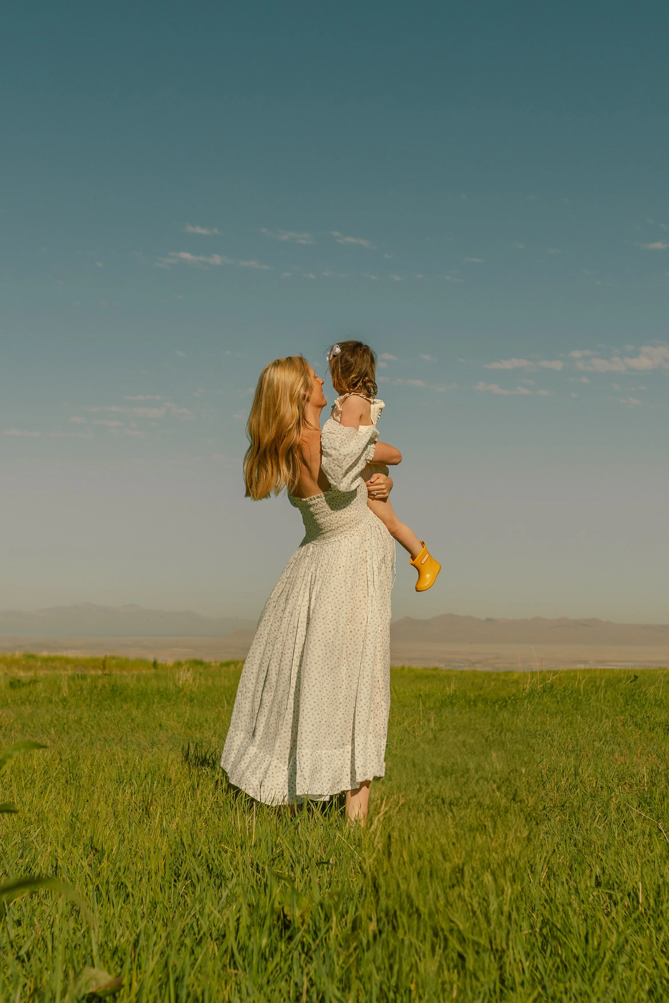 A woman in a long dress holding a child in a floral dress, standing on a grassy field with mountains in the distance and a clear blue sky above.