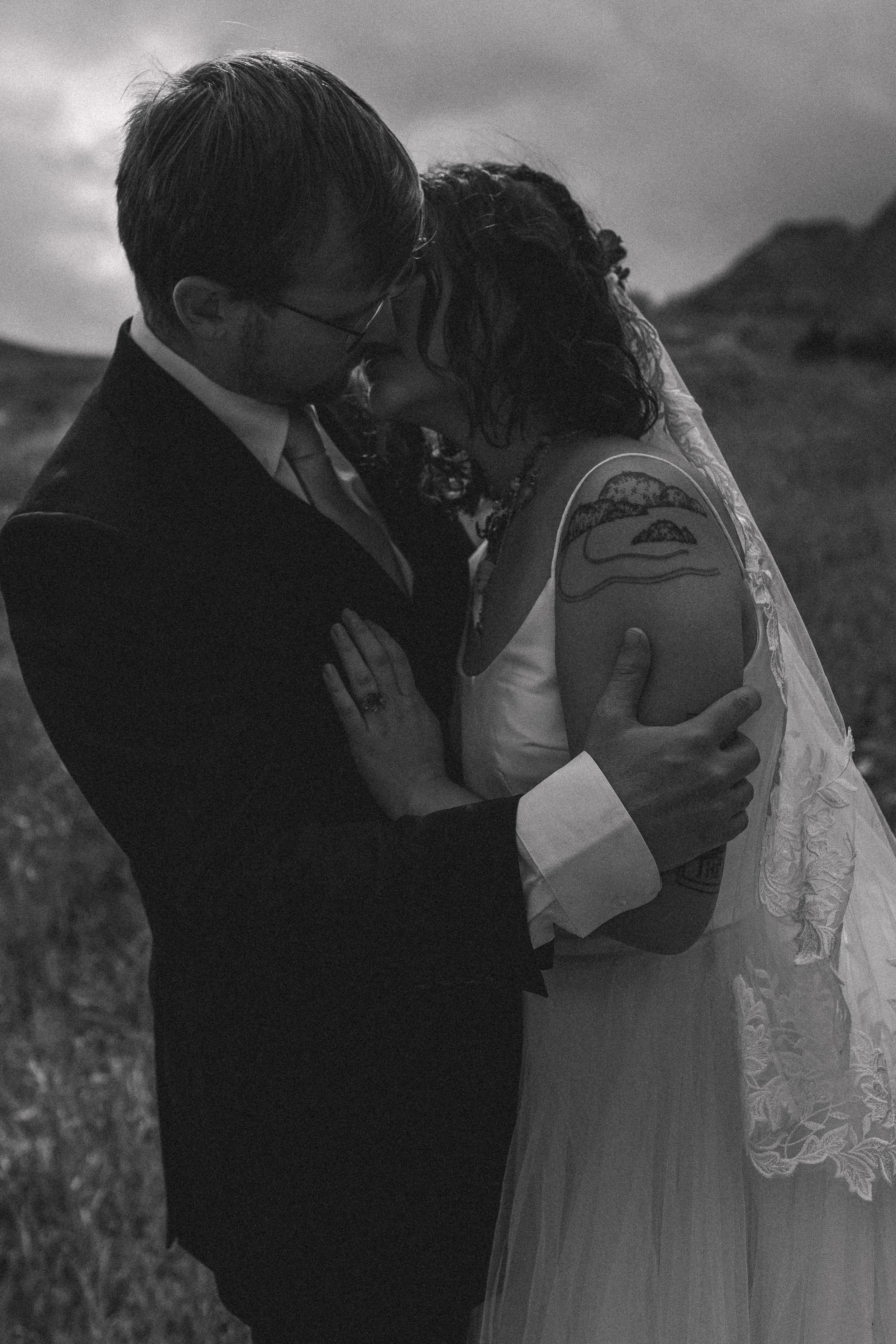 A black and white photograph of a couple in wedding attire embracing outdoors. The man wears glasses and a suit, while the woman has curly hair and a dress with lace details. They are holding each other closely with a mountain landscape in the backgr