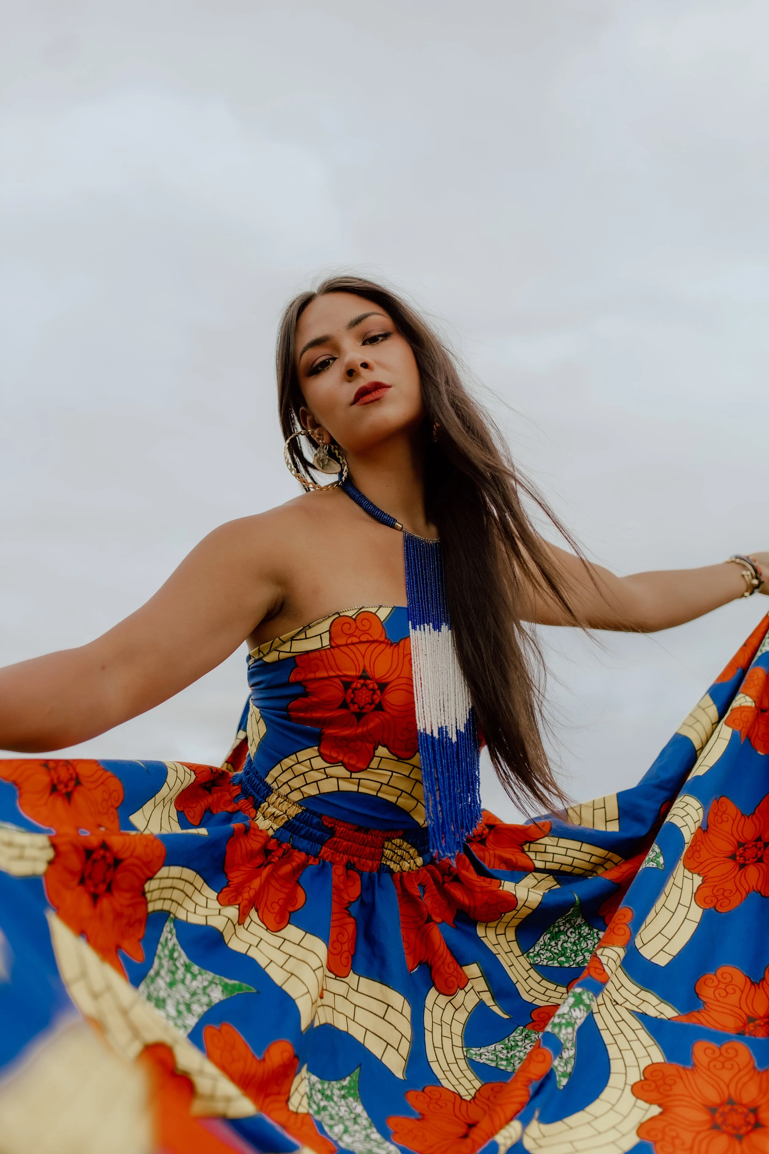 A woman dressed in a colorful, traditional African-style outfit with a long beaded necklace and large earrings, standing outdoors against a cloudy sky, with her arms outstretched and looking confidently at the camera.