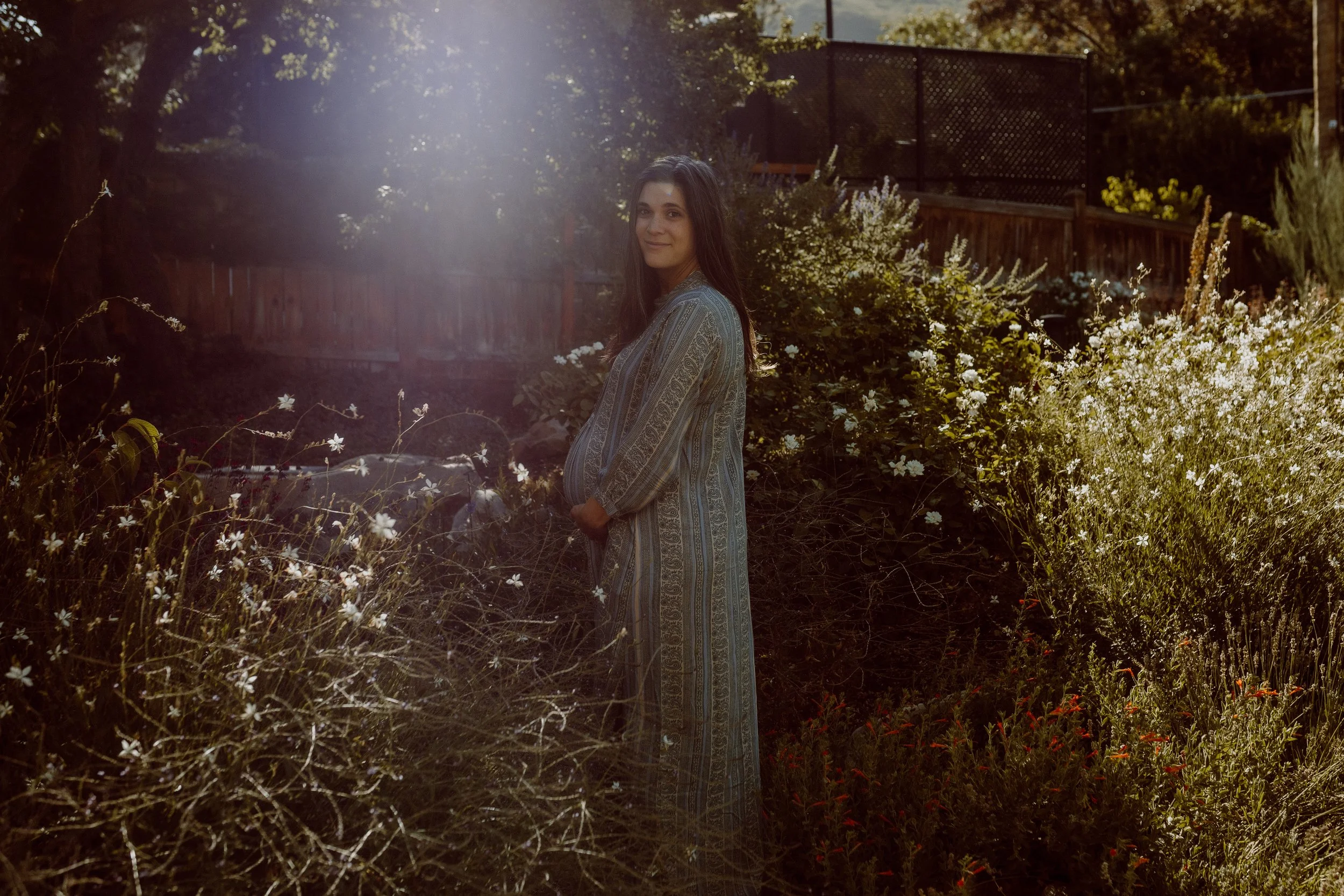 A woman with long brown hair wearing a patterned dress standing in a garden with white flowers, bushes, and trees during sunset.