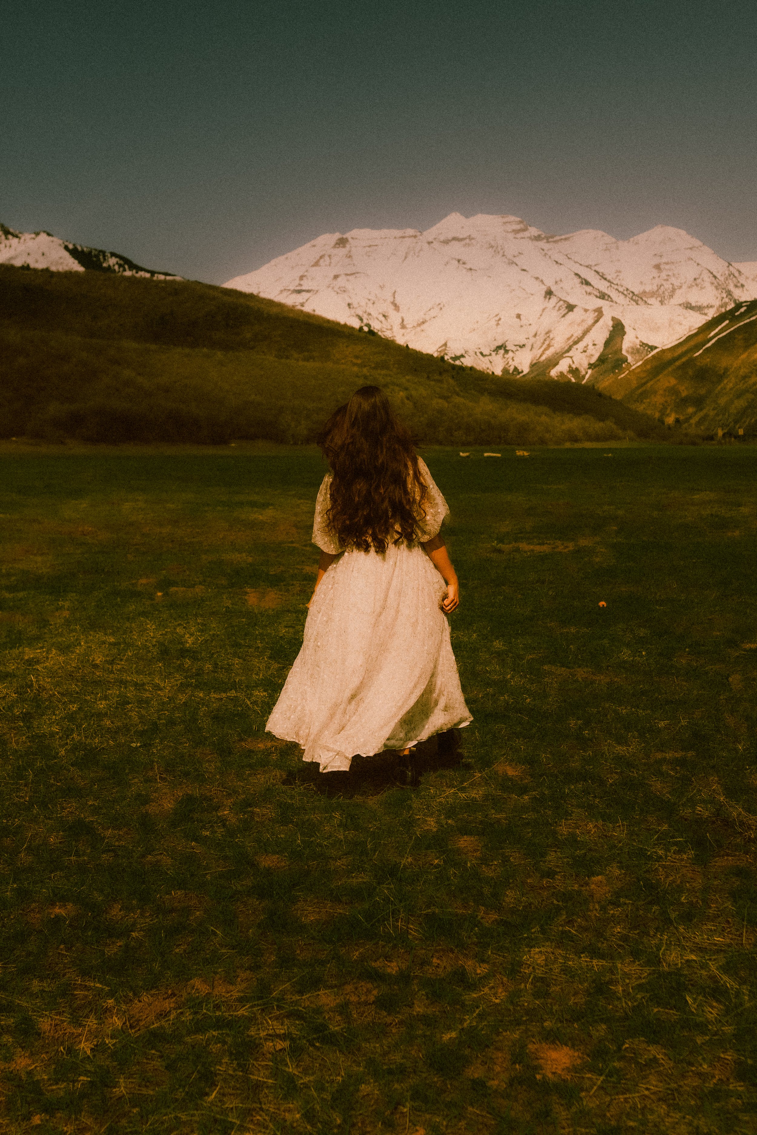 A woman with long, dark hair wearing a long, vintage-style white dress walking across a grassy field towards snow-capped mountains under a dark sky.
