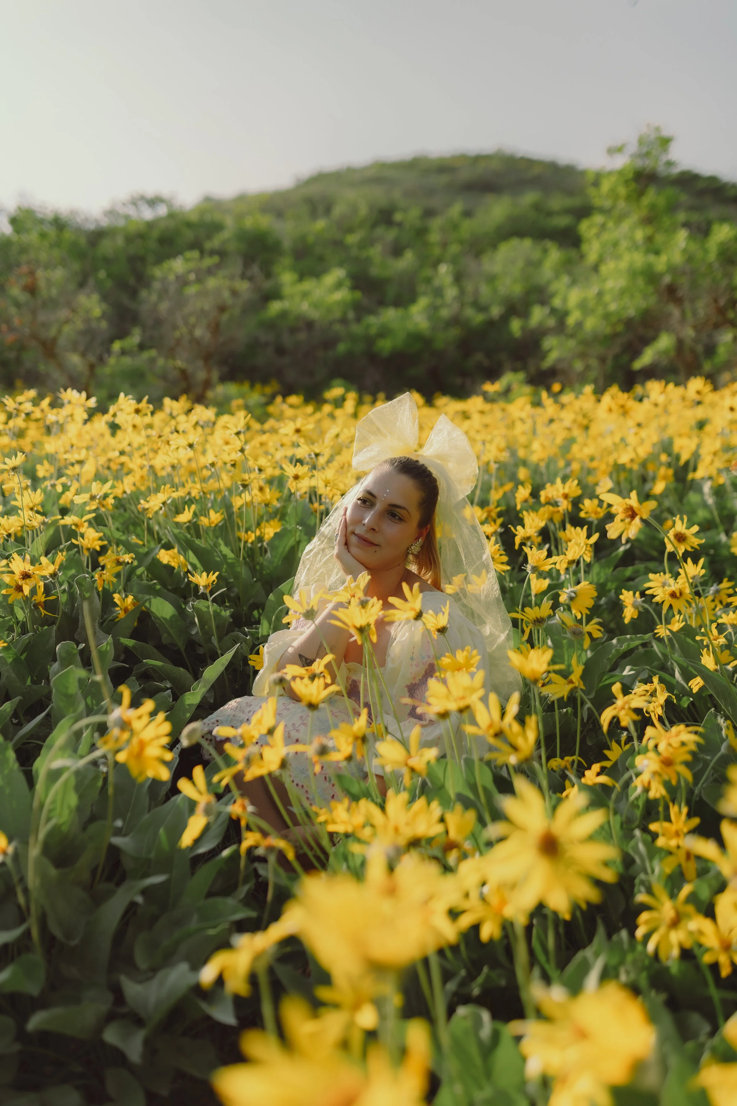 A woman in a yellow dress with a large yellow bow in her hair is sitting amidst a field of yellow flowers, with lush green trees and hills in the background.
