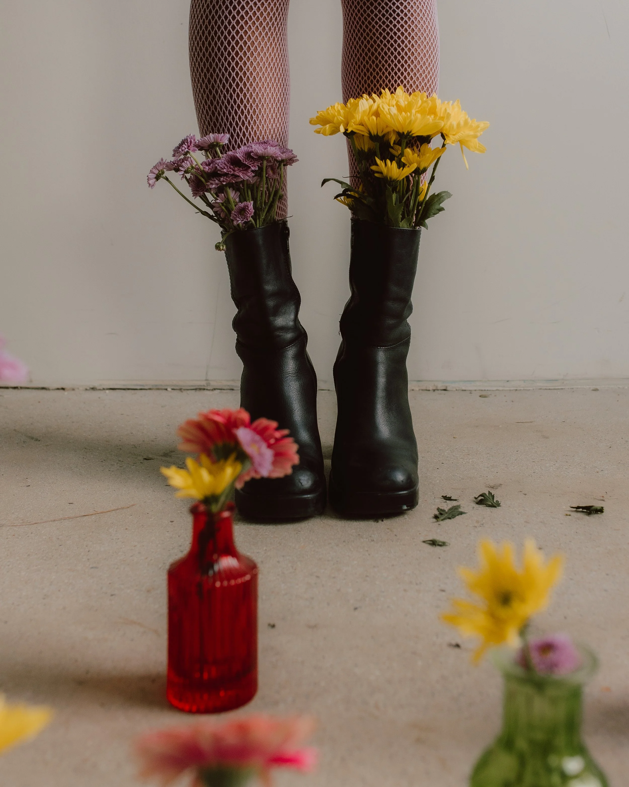 A person's legs wearing fishnet stockings and black boots, with flowers in the boots. The background is a plain wall and some scattered flowers on the floor.