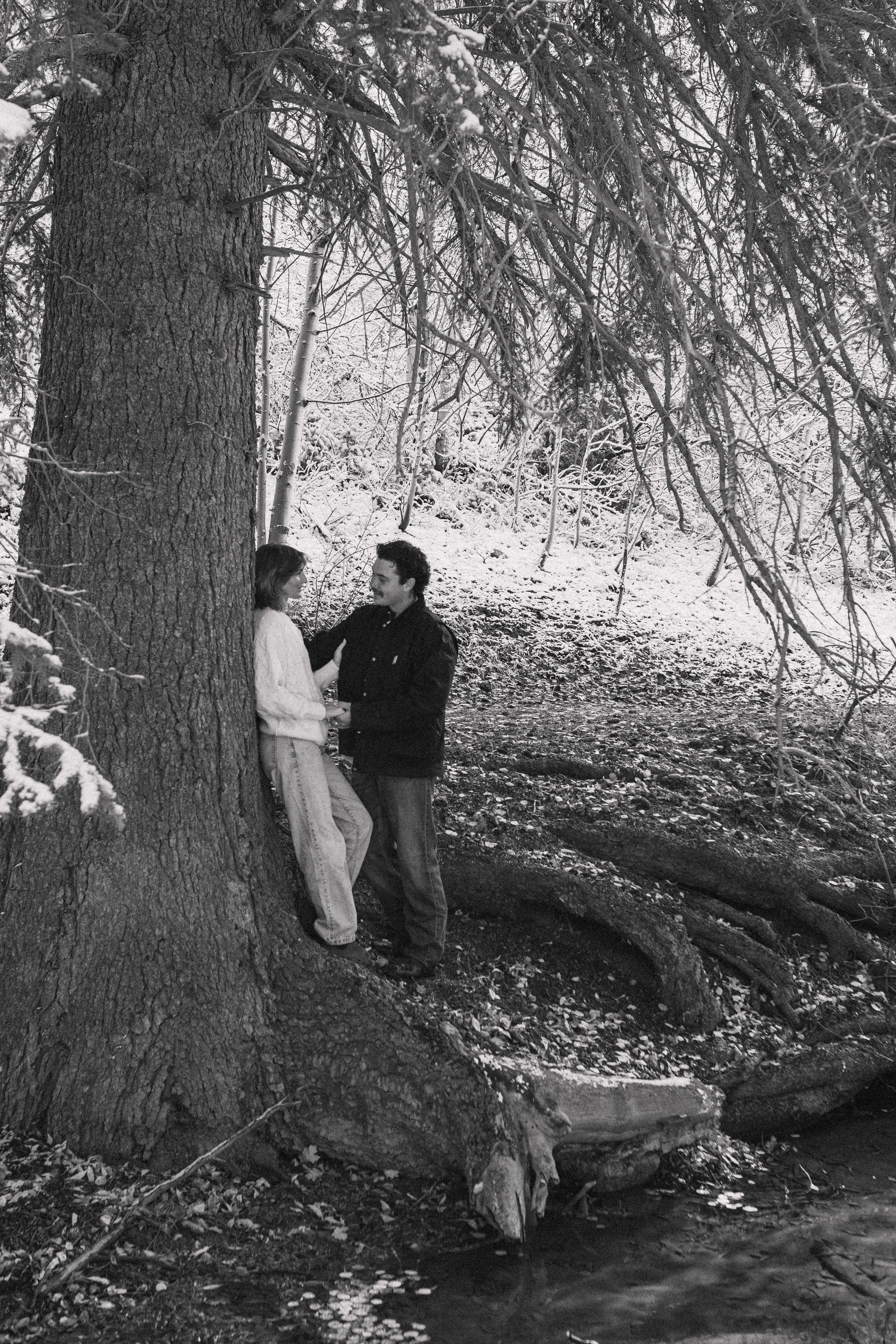 A black and white photo of a young couple standing by a large tree in a snowy forest. They are holding hands, looking at each other, and smiling. There is a fallen tree trunk and a small stream in the background.