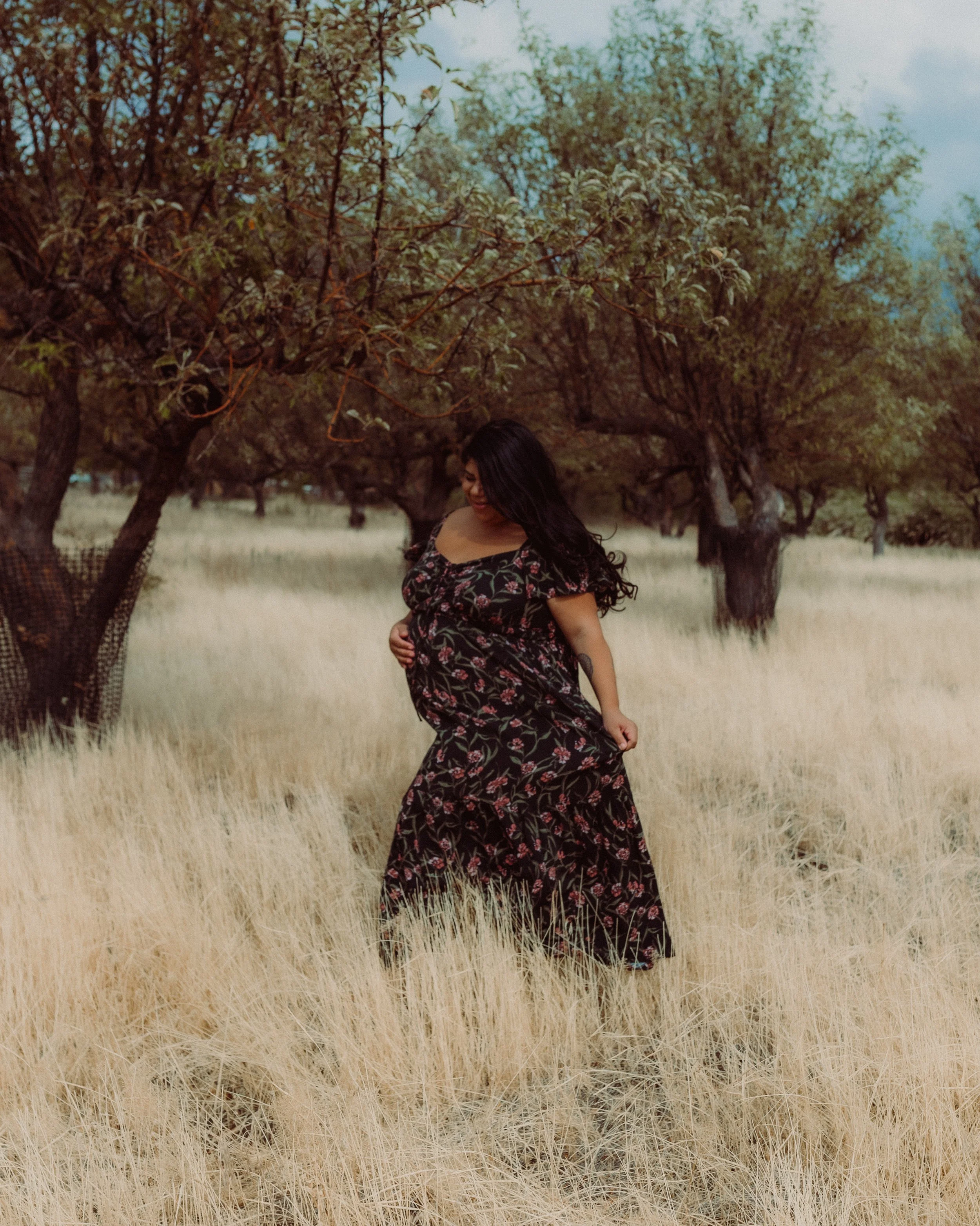 A woman wearing a floral dress stands in a field of tall wheat grass with a line of trees with green and brown leaves behind her, under a partly cloudy sky.
