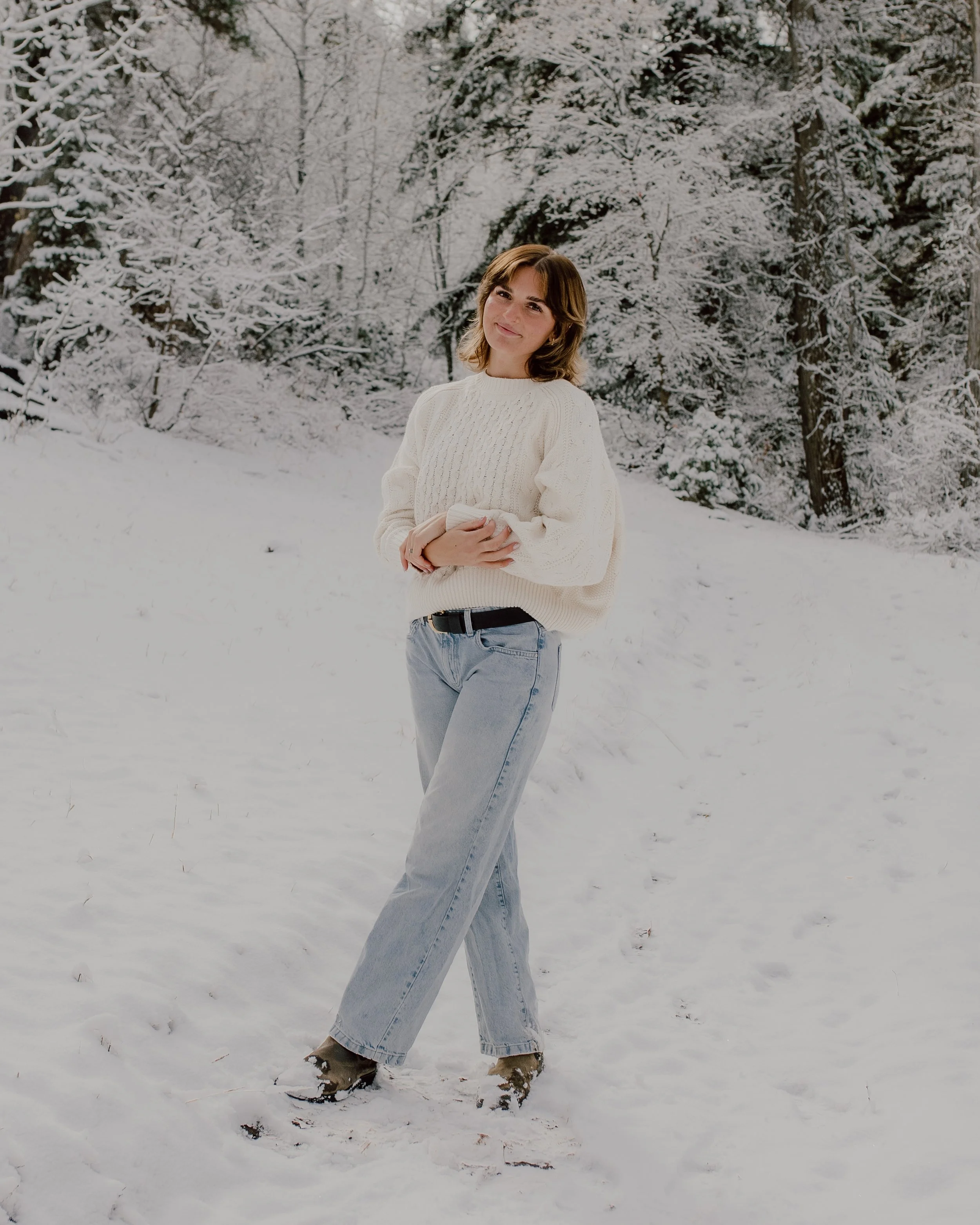 A young woman with shoulder-length brown hair and a light complexion stands in a snowy forest. She is smiling and looking at the camera, wearing a cream-colored sweater and light blue jeans. Snow covers the ground and trees in the background.