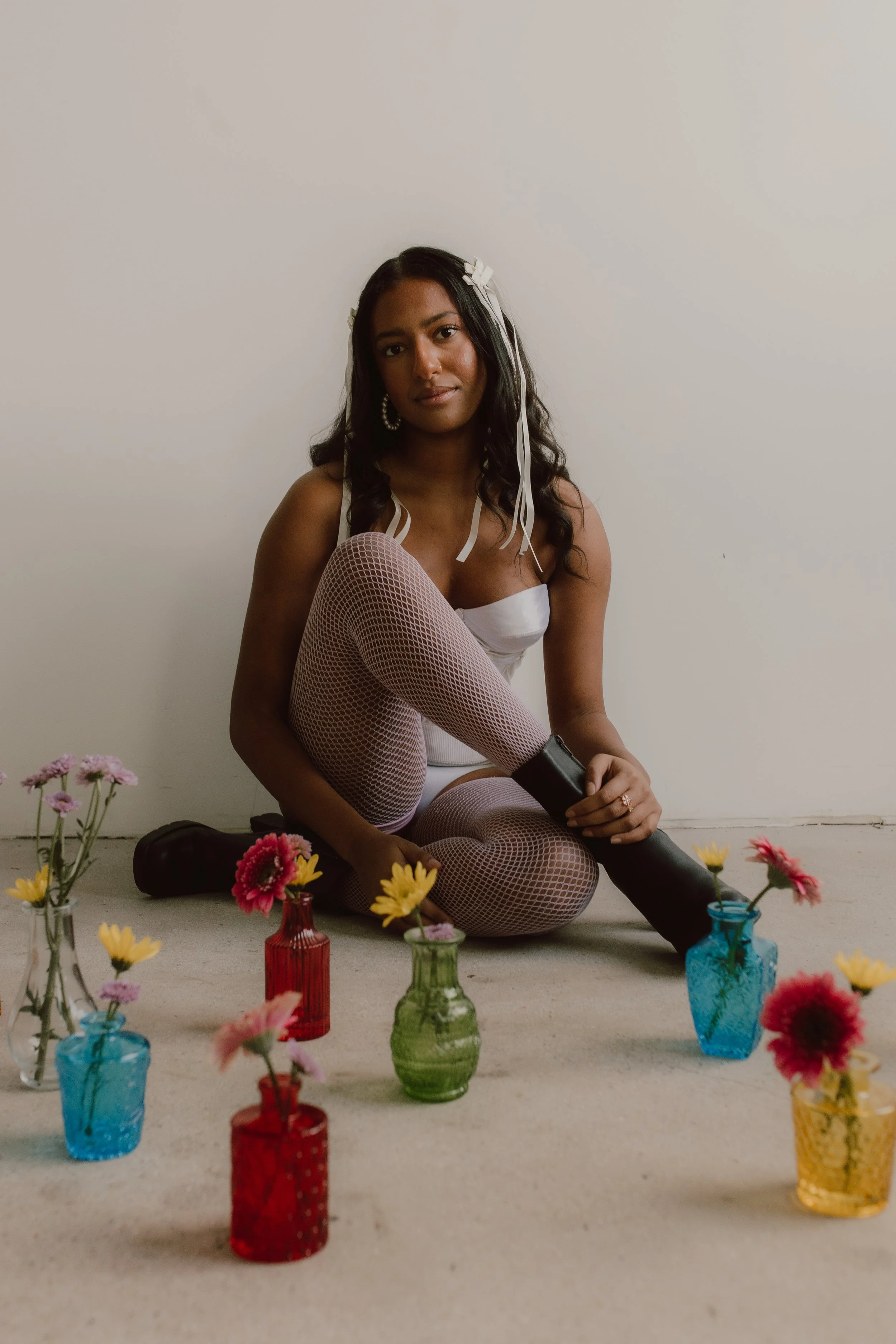 A woman with long dark hair sitting on the floor in front of a plain white background, surrounded by colorful vases with flowers.