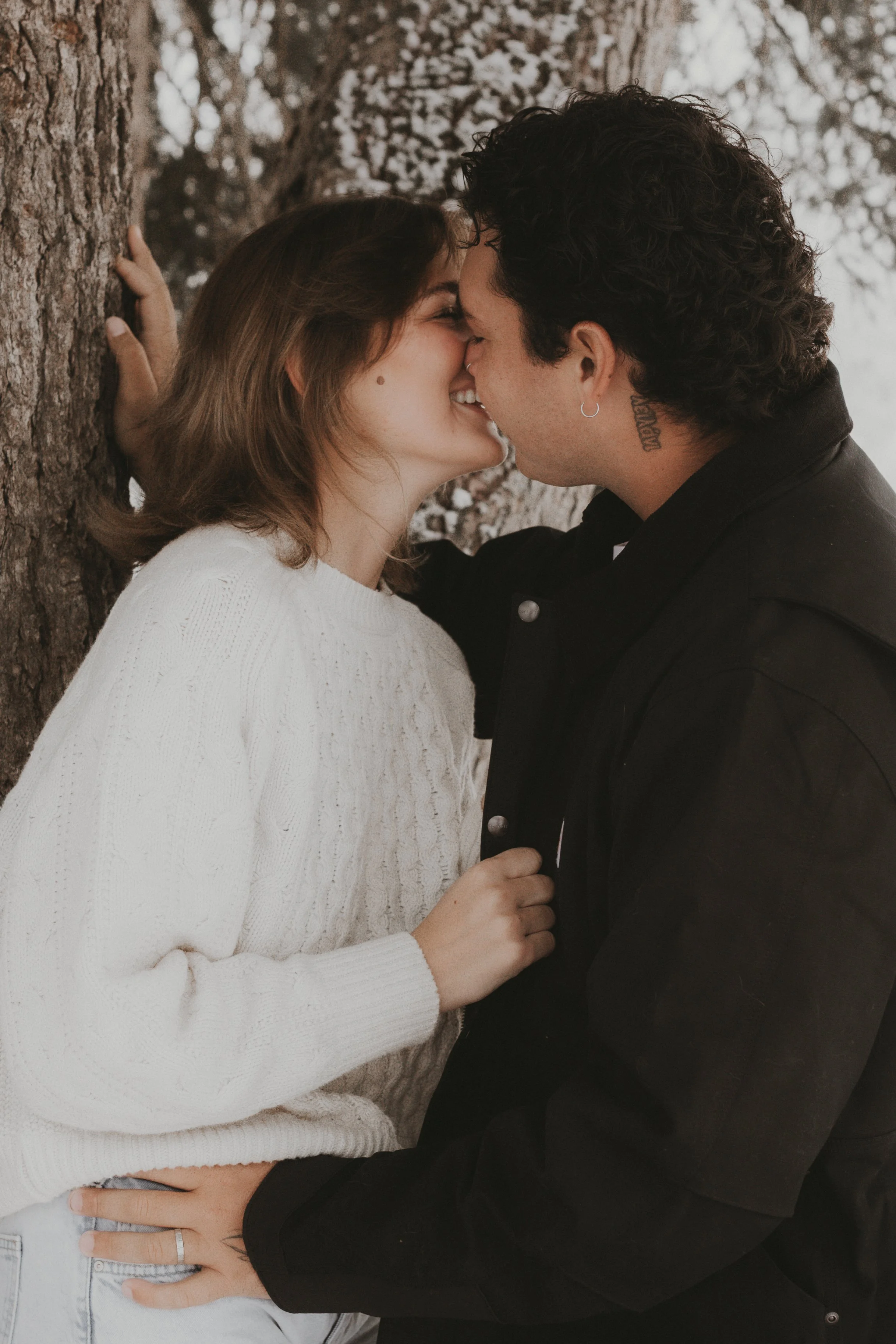 A close-up of a couple about to kiss, with their faces touching and eyes closed. The woman has medium-length brown hair, a beauty mark on her cheek, and is wearing a white sweater. The man has curly dark hair, a hoop earring, a tattoo on his neck, an
