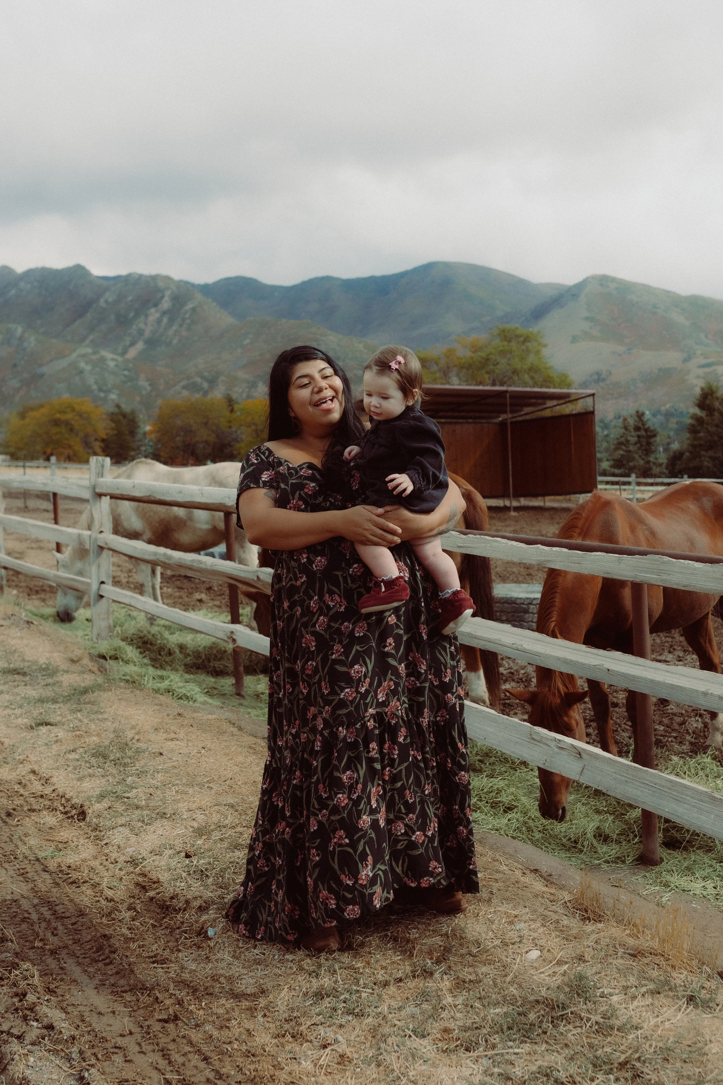A woman holding a small child at a horse farm with mountains in the background. The woman is wearing a long black floral dress, and the child is wearing a black jacket and red shoes. Horses are visible behind a wooden fence.
