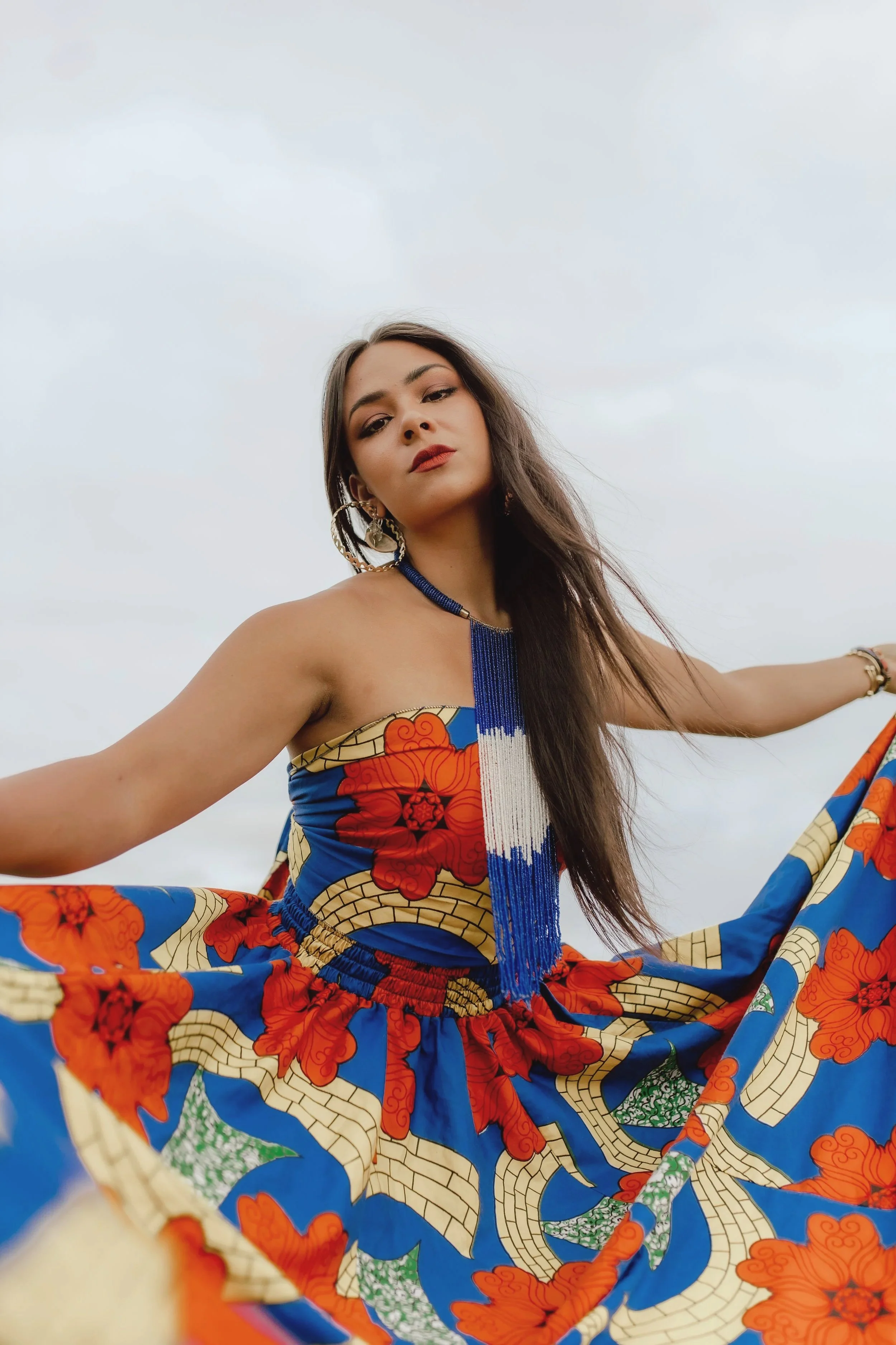 Young woman in colorful, patterned dress with blue, red, orange, and green designs, wearing large earrings, standing outside against cloudy sky, with arms extended outward.