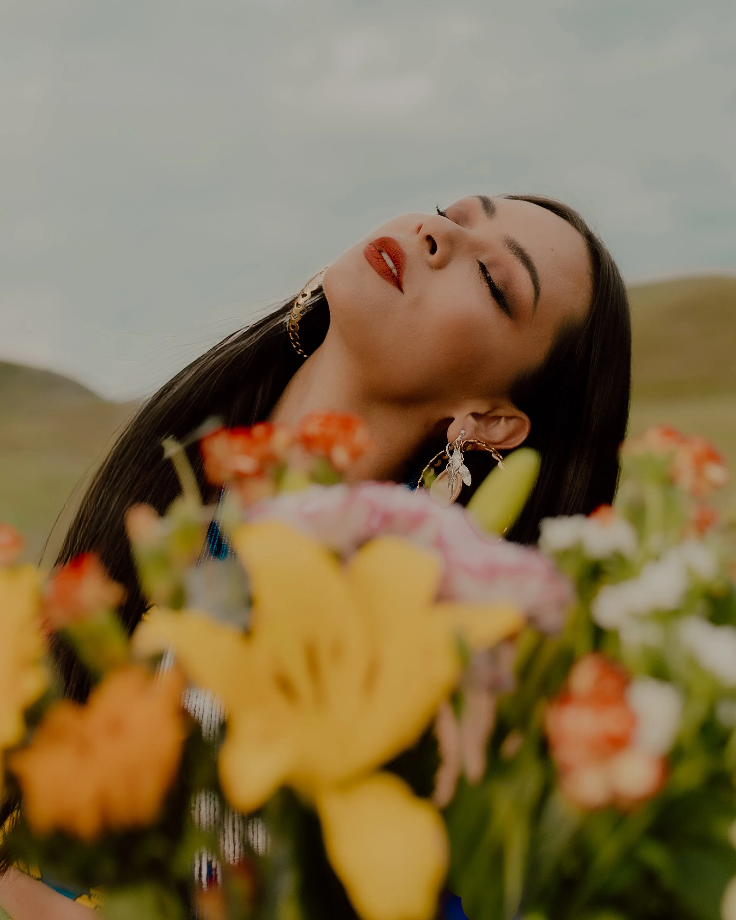 A woman with long dark hair and makeup, including red lipstick and eye makeup, is outdoors during daytime with a cloudy sky in the background. She is wearing earrings and a chain necklace, and is surrounded by colorful flowers.