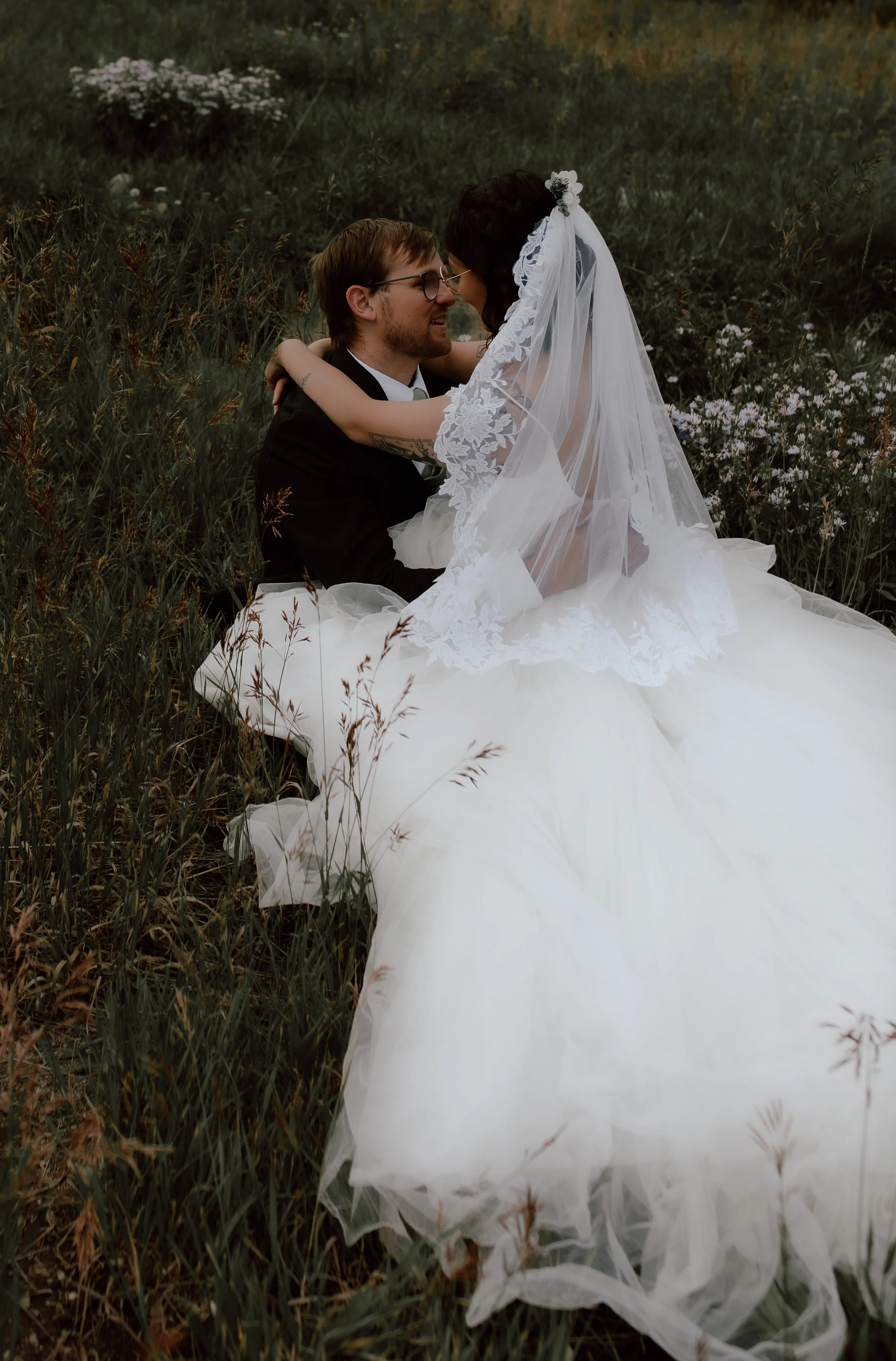 A bride and groom sitting in a field, embracing, with the bride wearing a lace veil and a white wedding gown, and the groom in a black suit. The surrounding area has tall grass and wildflowers.