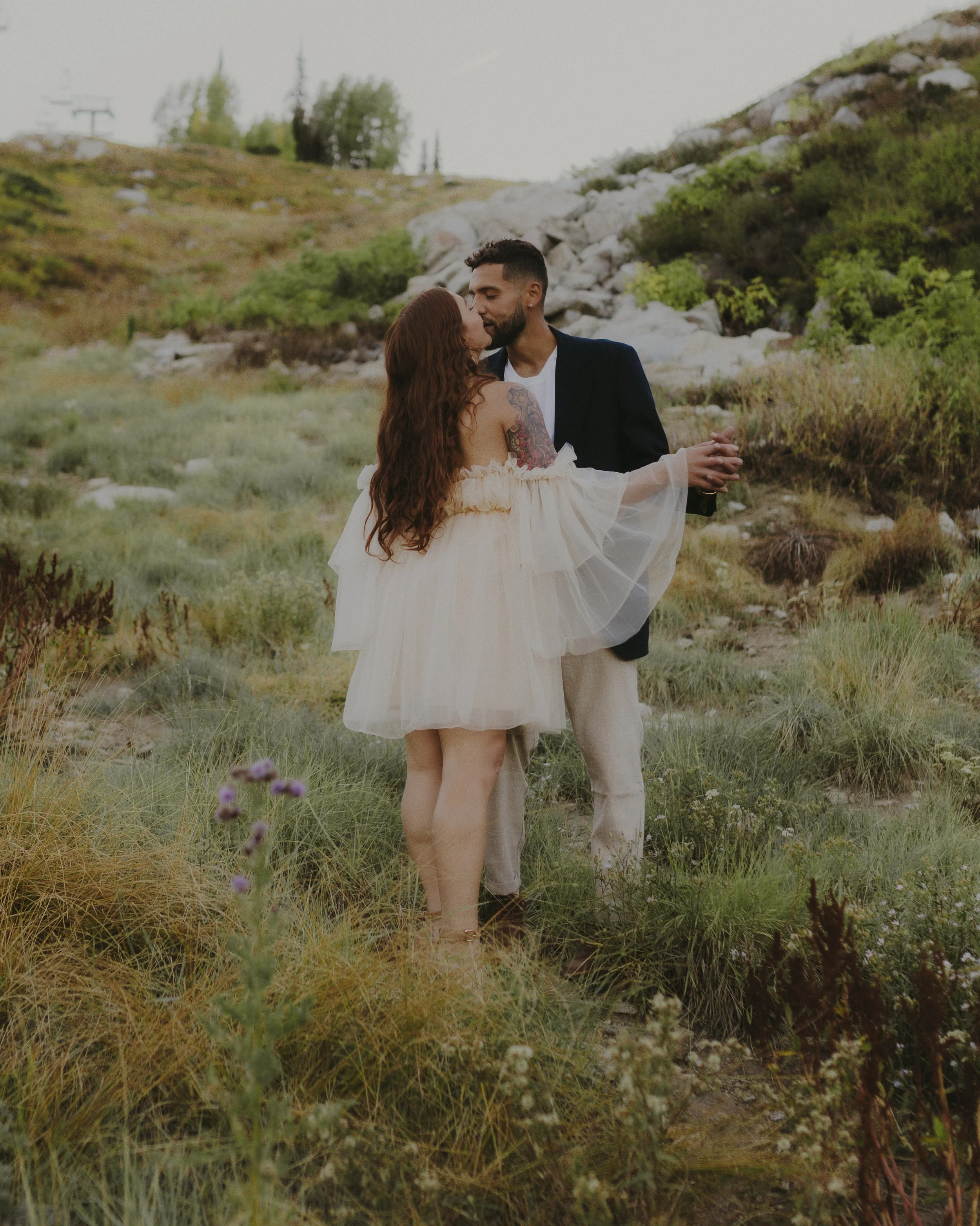 A couple dancing outdoors in a grassy area with hills, rocks, and trees in the background. The woman has long red hair, a tattoo on her arm, and is wearing an off-shoulder white dress. The man has short dark hair, a beard, and is dressed in a blazer 