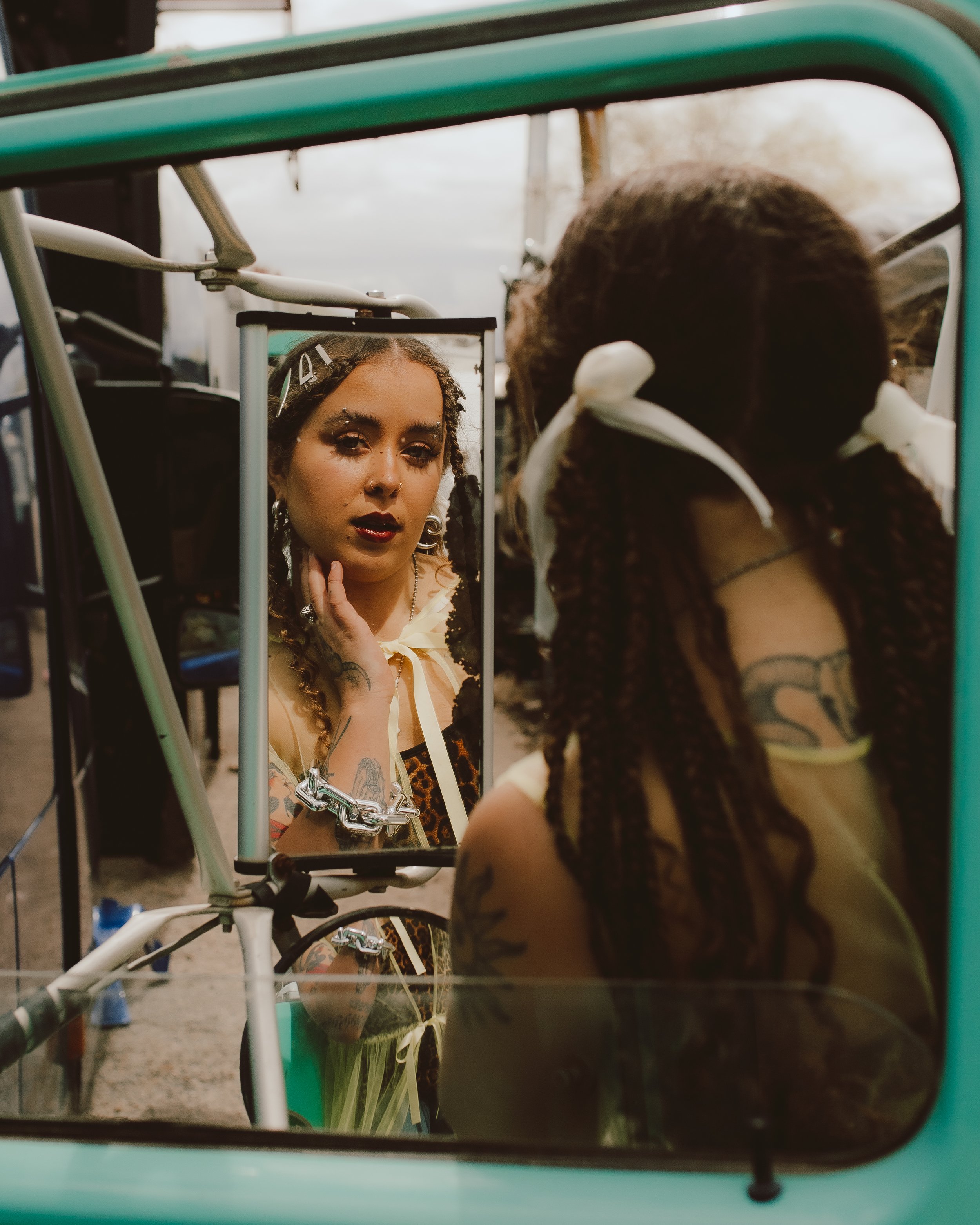A woman with tattoos on her arm and makeup, looking into a mirror attached to a piece of gym equipment, with her reflection visible in the mirror.