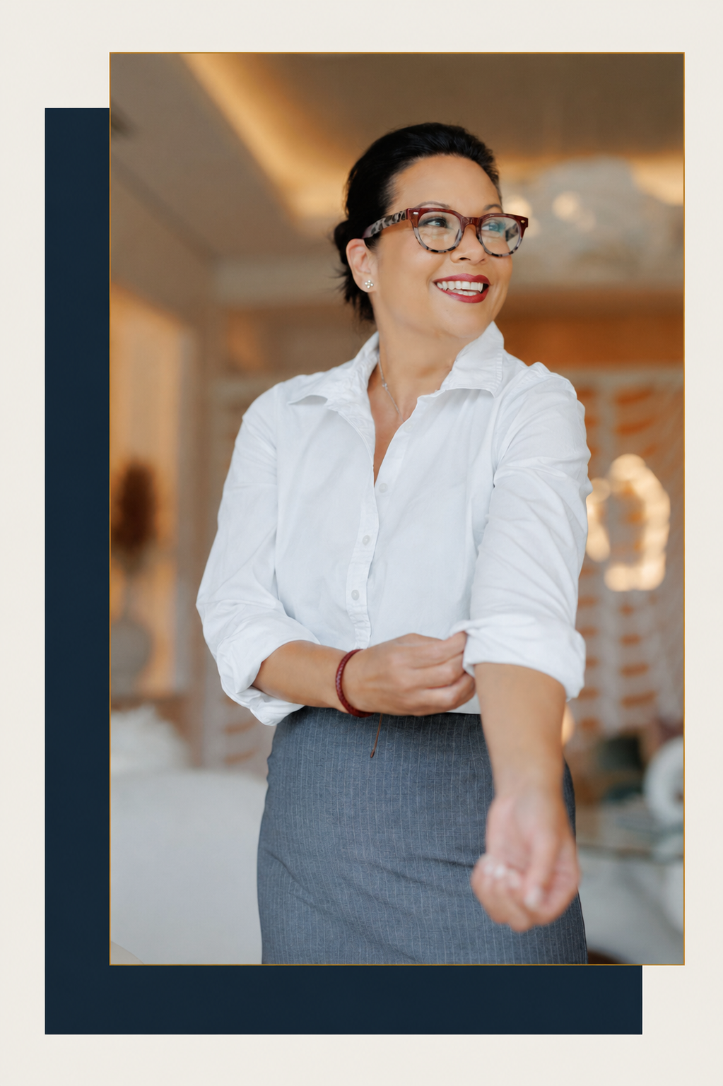 A woman with short dark hair, glasses, and wearing a white button-up shirt and gray skirt, smiling and rolling up her long sleeve
