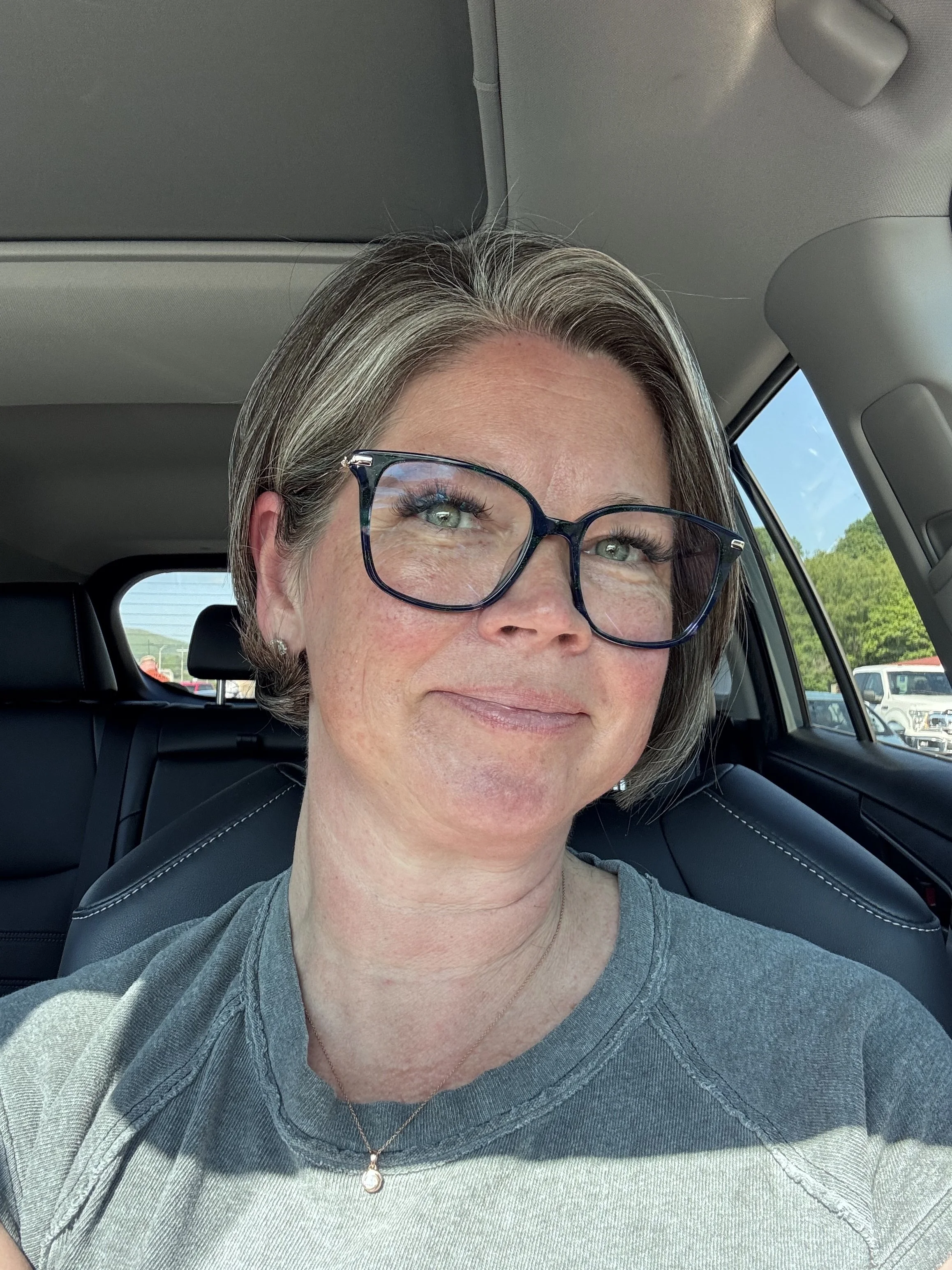 Close-up of a middle-aged woman with short gray hair, glasses, and a light gray T-shirt, sitting in a car vehicle with a view of blue sky and green trees outside.