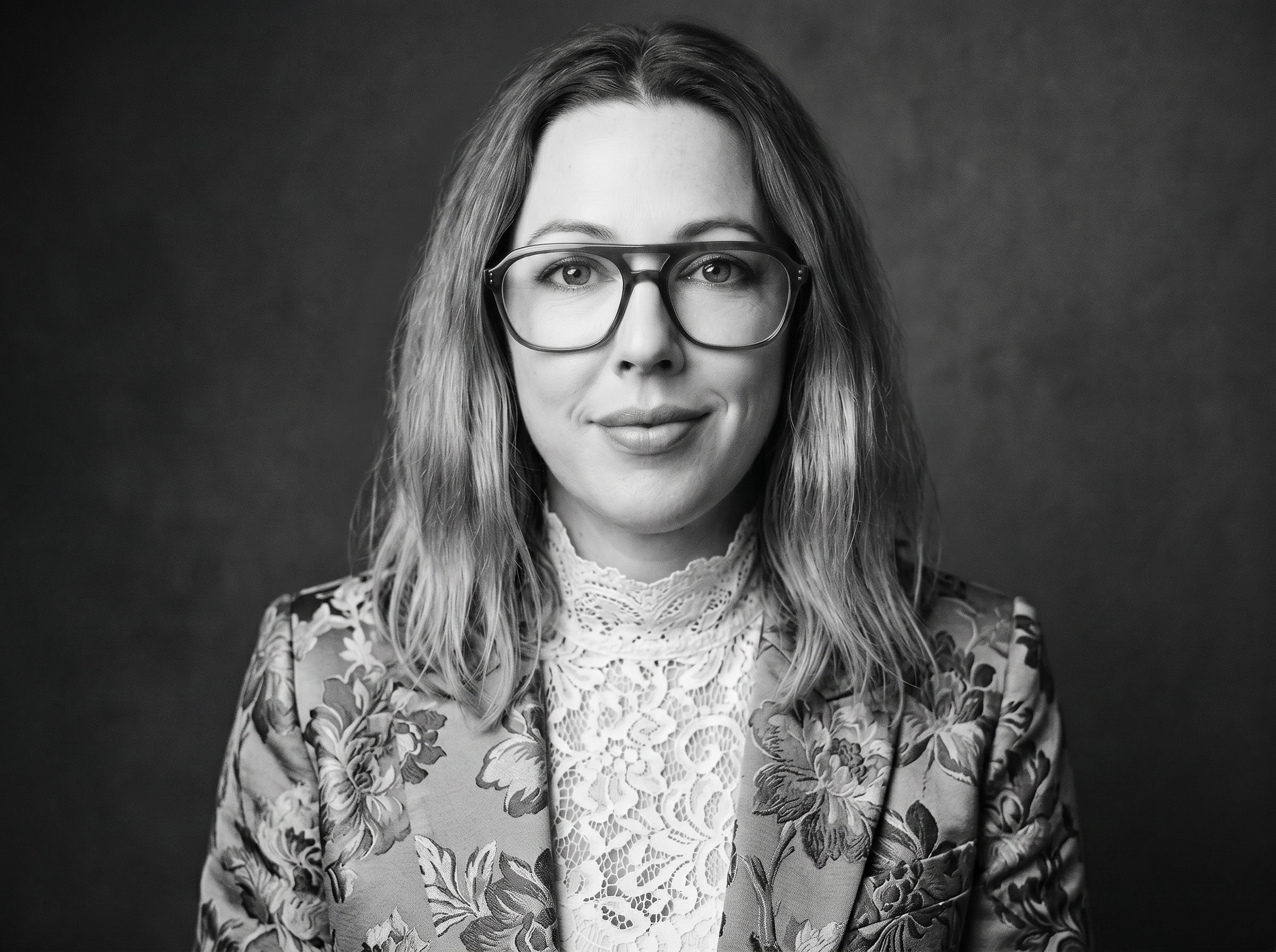 Black and white portrait of a woman with long wavy hair wearing large glasses and a floral lace top, smiling slightly against a plain dark background.