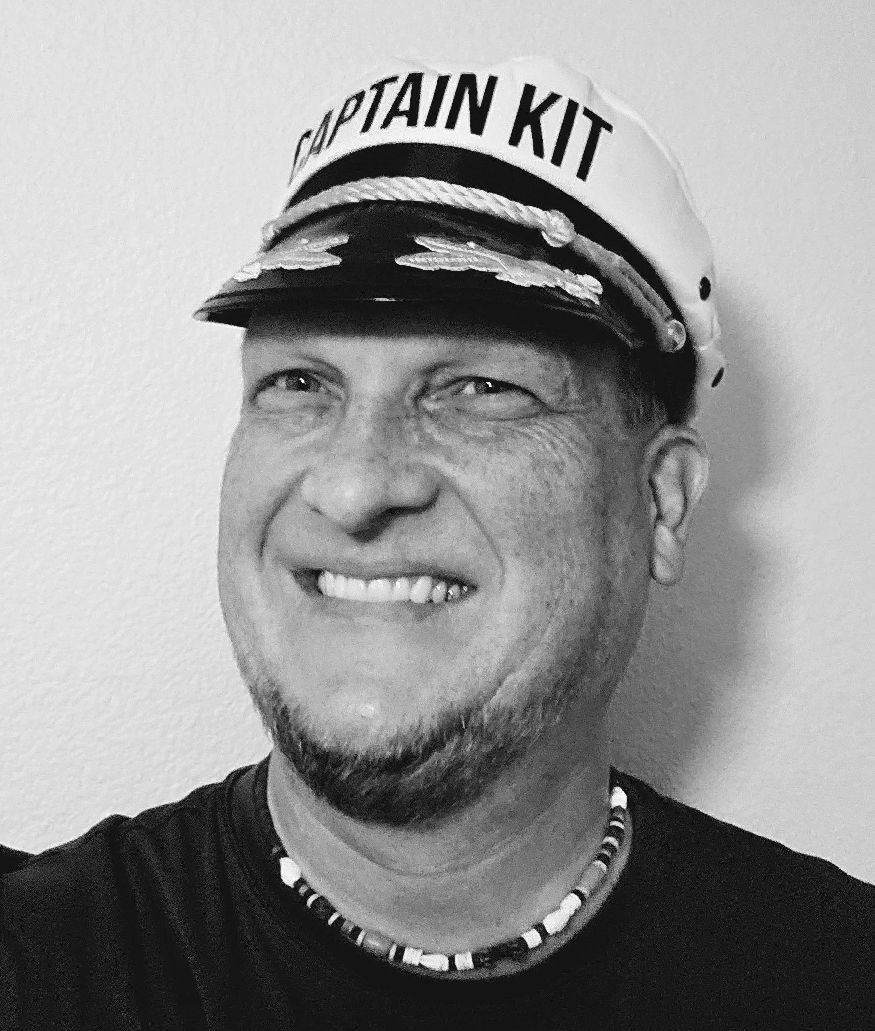 A black and white close-up photograph of a smiling man wearing a cap labeled 'Captain Kit' and a beaded necklace.
