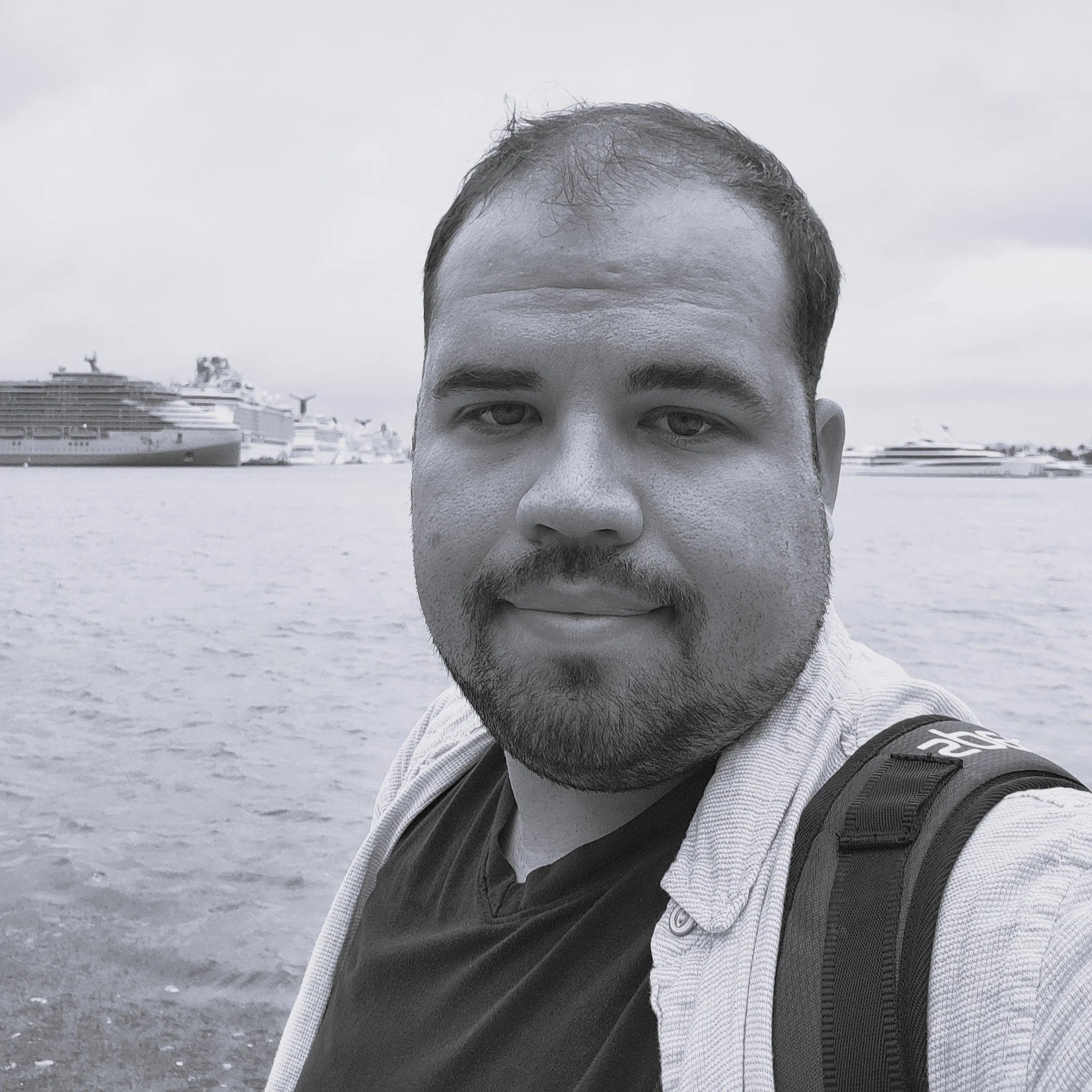 Close-up of a man's face from the nose up, with the background showing a harbor with large cruise ships and an overcast sky.