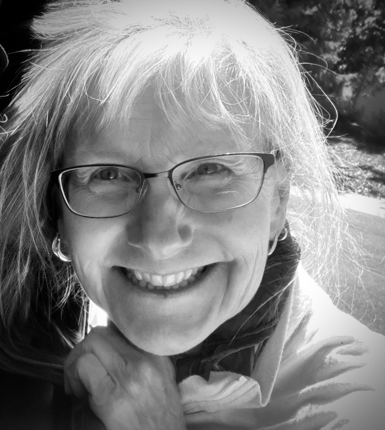 A smiling woman with glasses, shoulder-length hair, earrings, and a scarf, outdoors in sunlight.