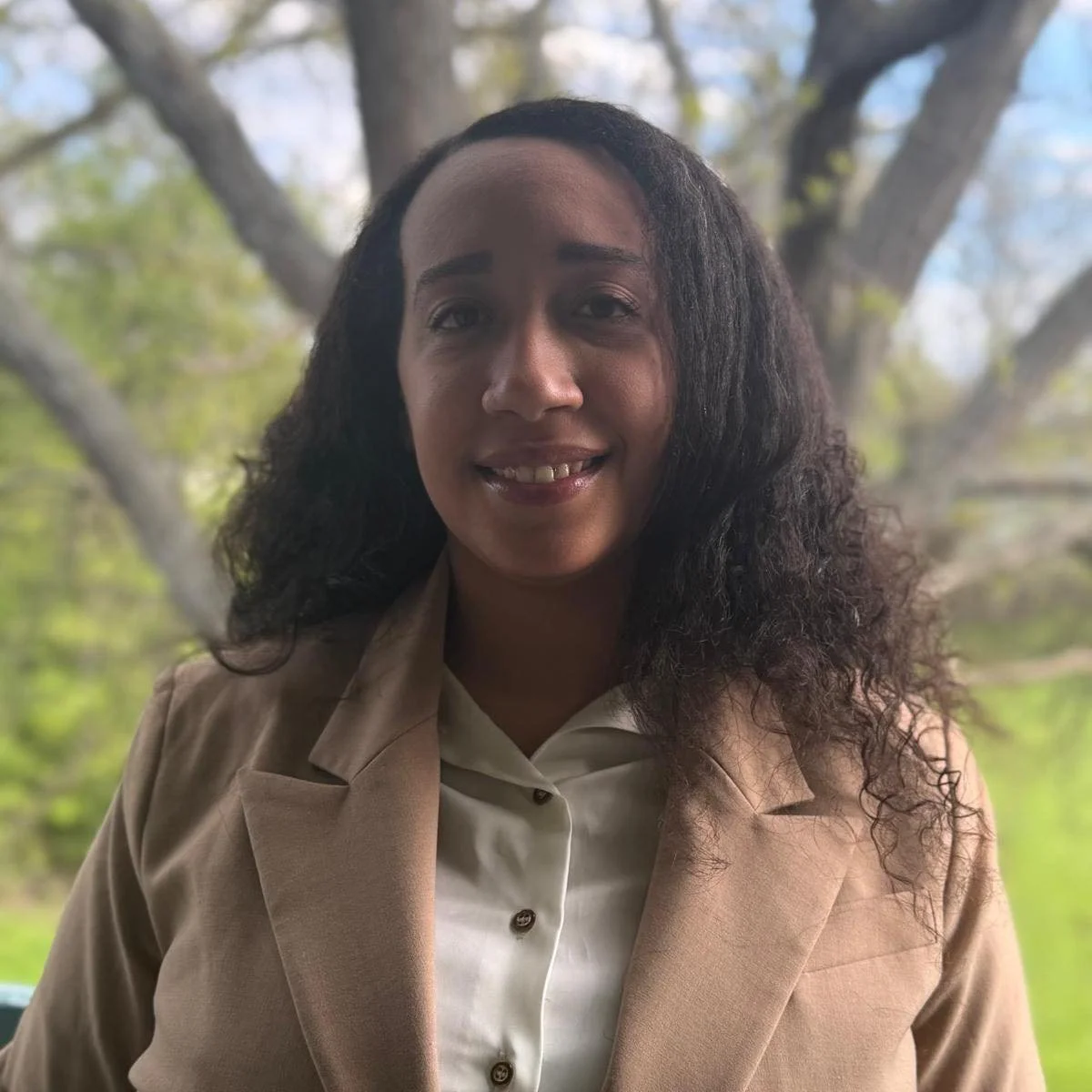 A woman with curly black hair smiling outdoors, wearing a beige blazer and white blouse, with trees and a cloudy sky in the background.