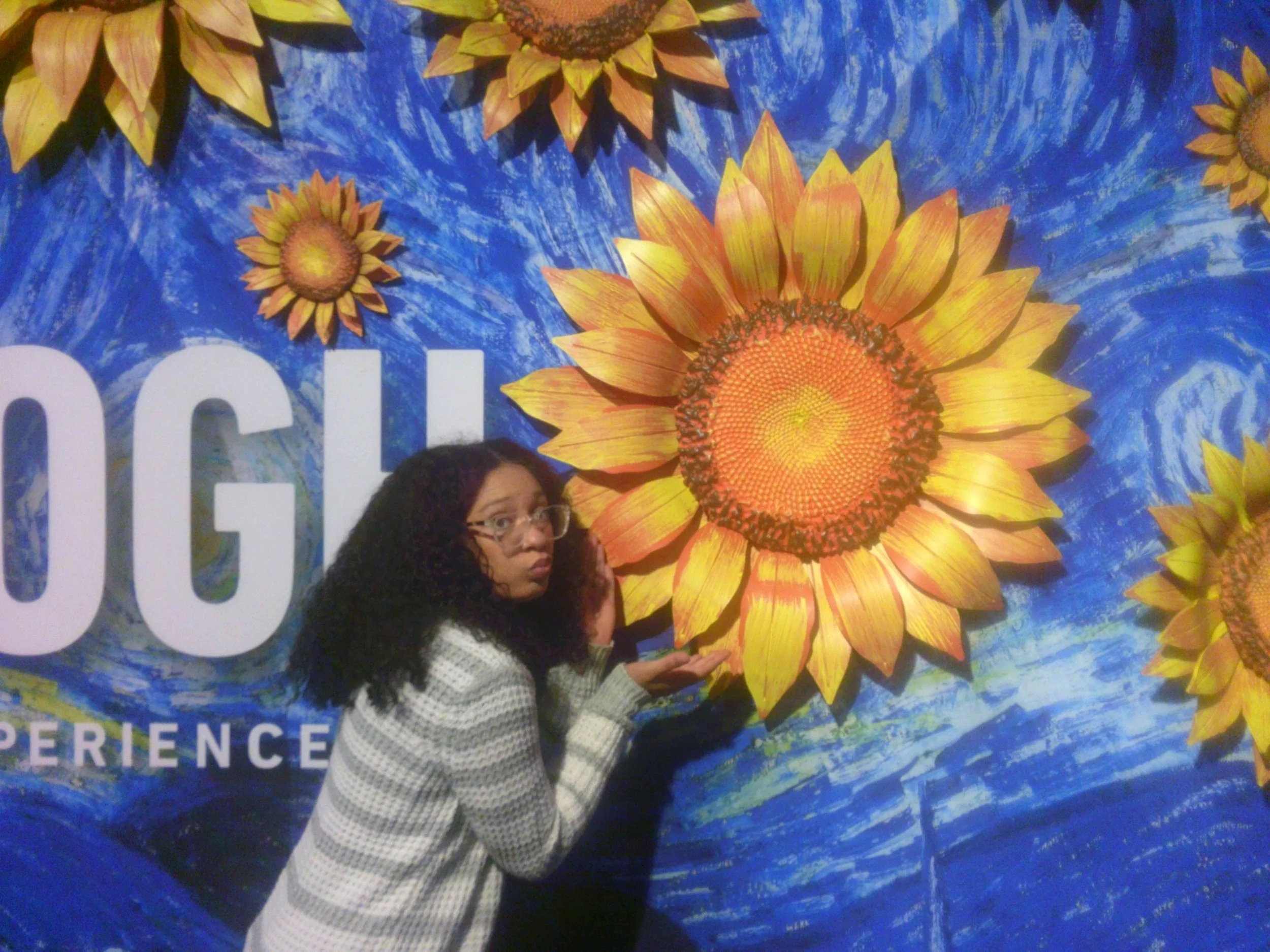 A woman with curly black hair and glasses wearing a black and white checkered sweater, standing in front of a colorful mural of sunflowers and blue sky, partially covering the mural.