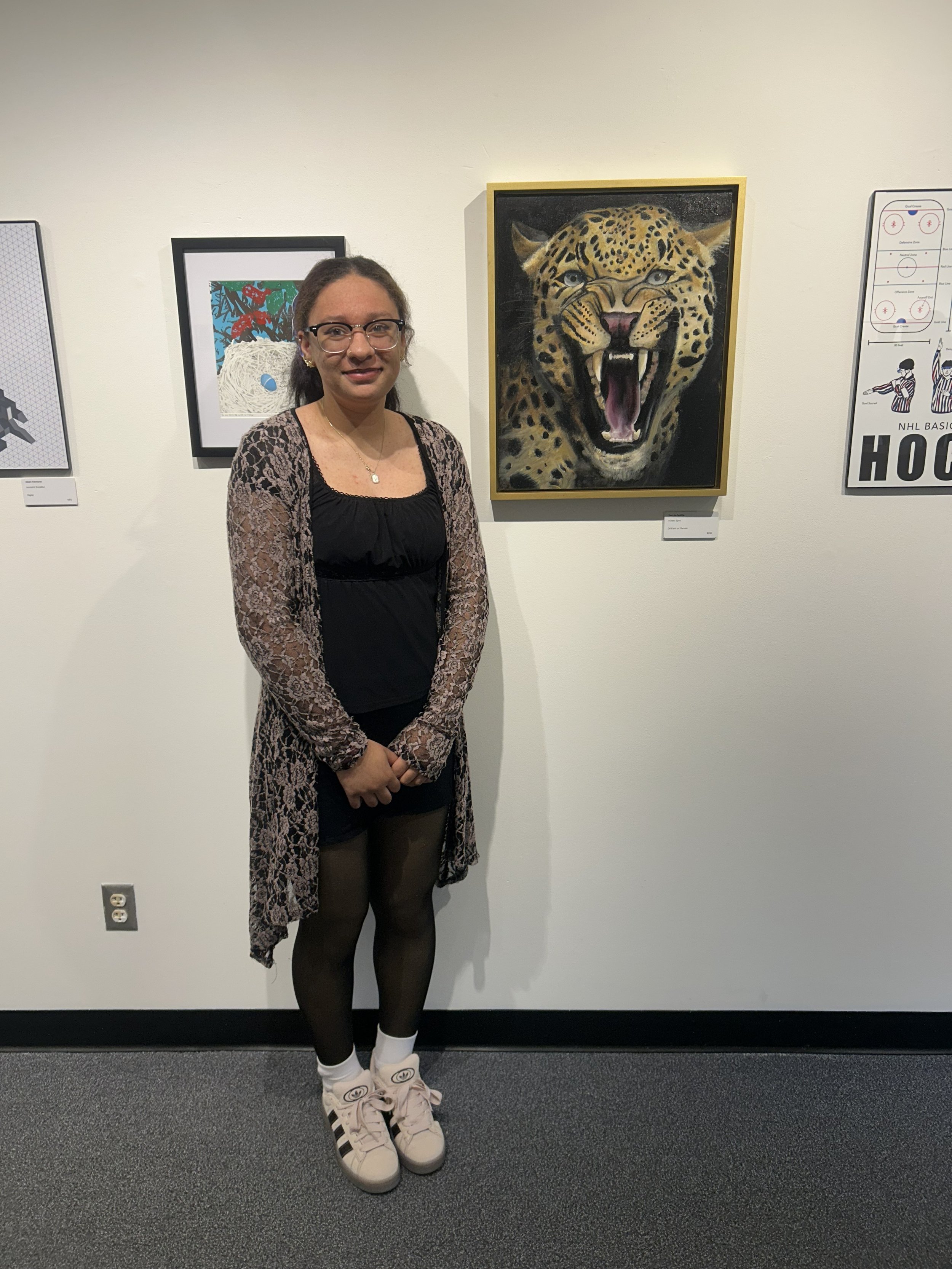 A woman standing in an art gallery, wearing black tights, white sneakers with black stripes, a black dress, and a lace cardigan, posing in front of a painting of a roaring jaguar on a white wall.