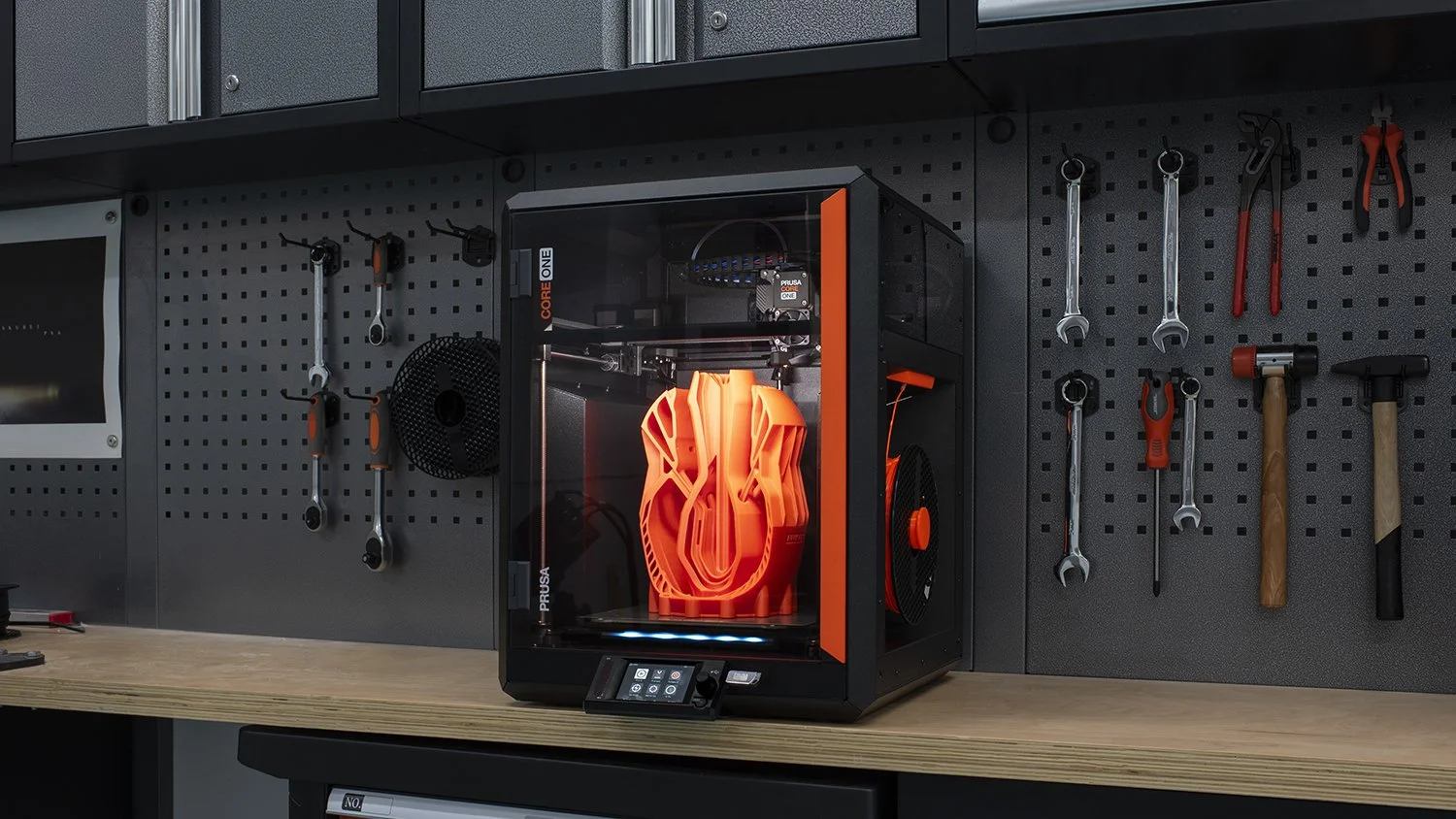 A 3D printer with a black exterior and a glass door shows an orange 3D printed object inside. The printer is on a wooden workbench with a pegboard behind it holding various tools like wrenches, screwdrivers, pliers, and hammers.