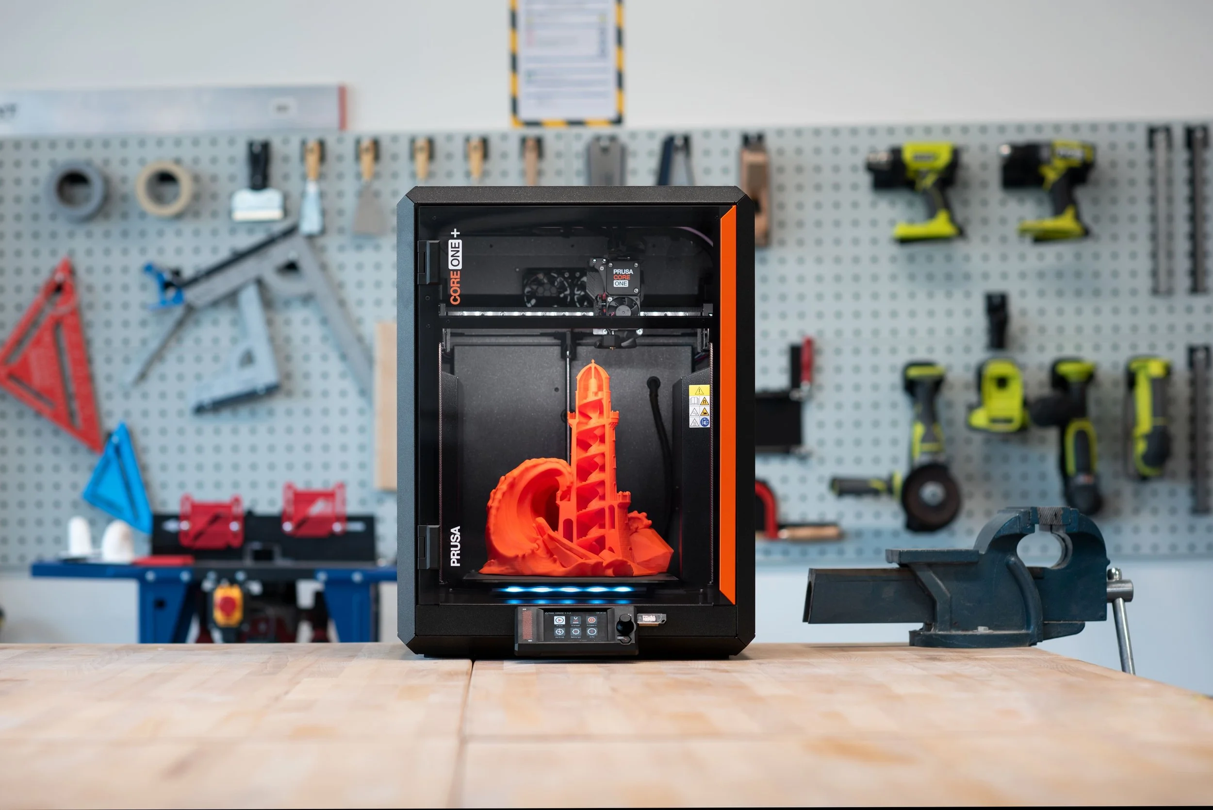 A 3D printer printing an orange structure in a workshop with tools and supplies on a pegboard in the background.