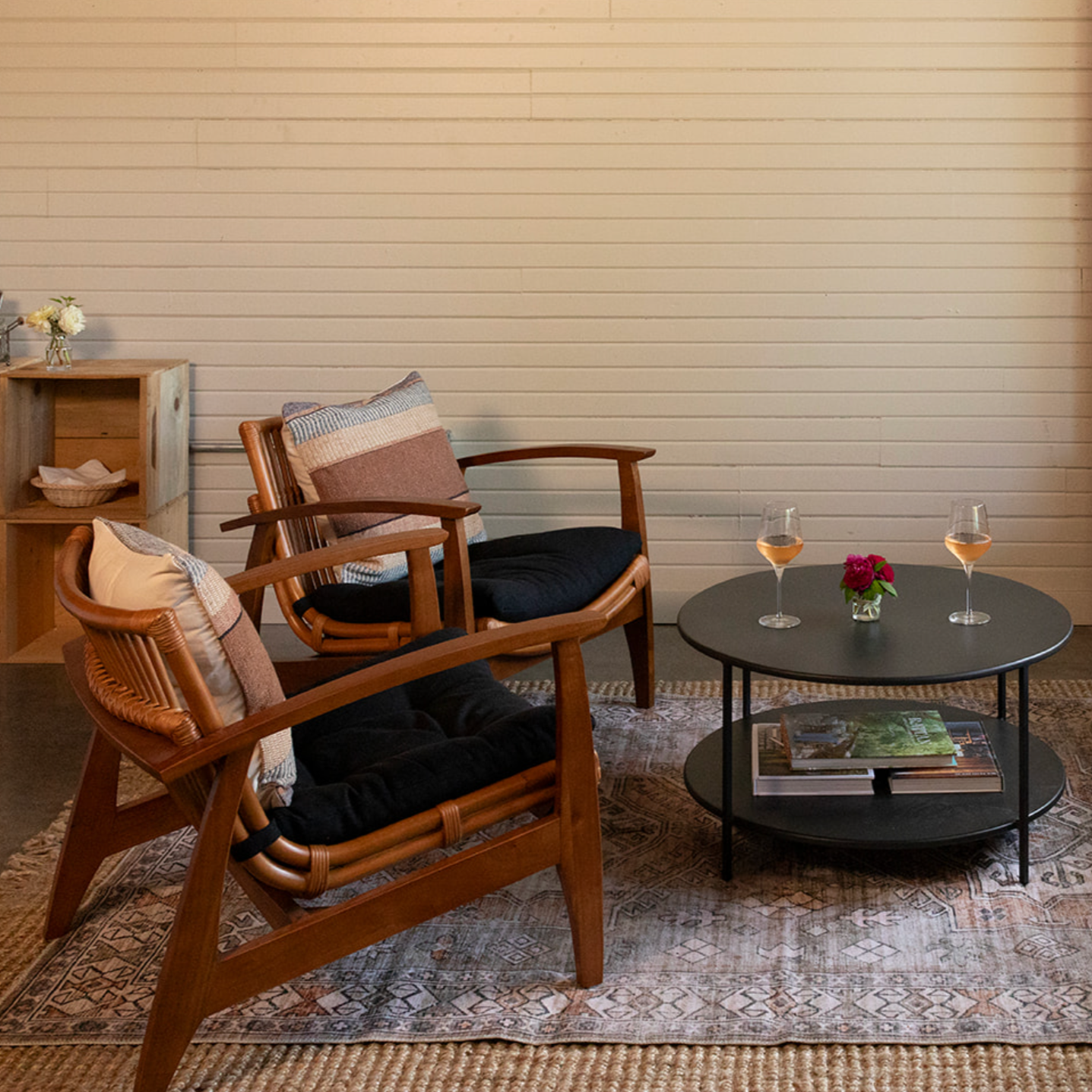 Living room with two wooden armchairs with black cushions and patterned pillows, a black round coffee table with two glasses of rosé wine, a small vase with pink flowers, and books underneath, a wooden bookshelf, and a beige brick wall.