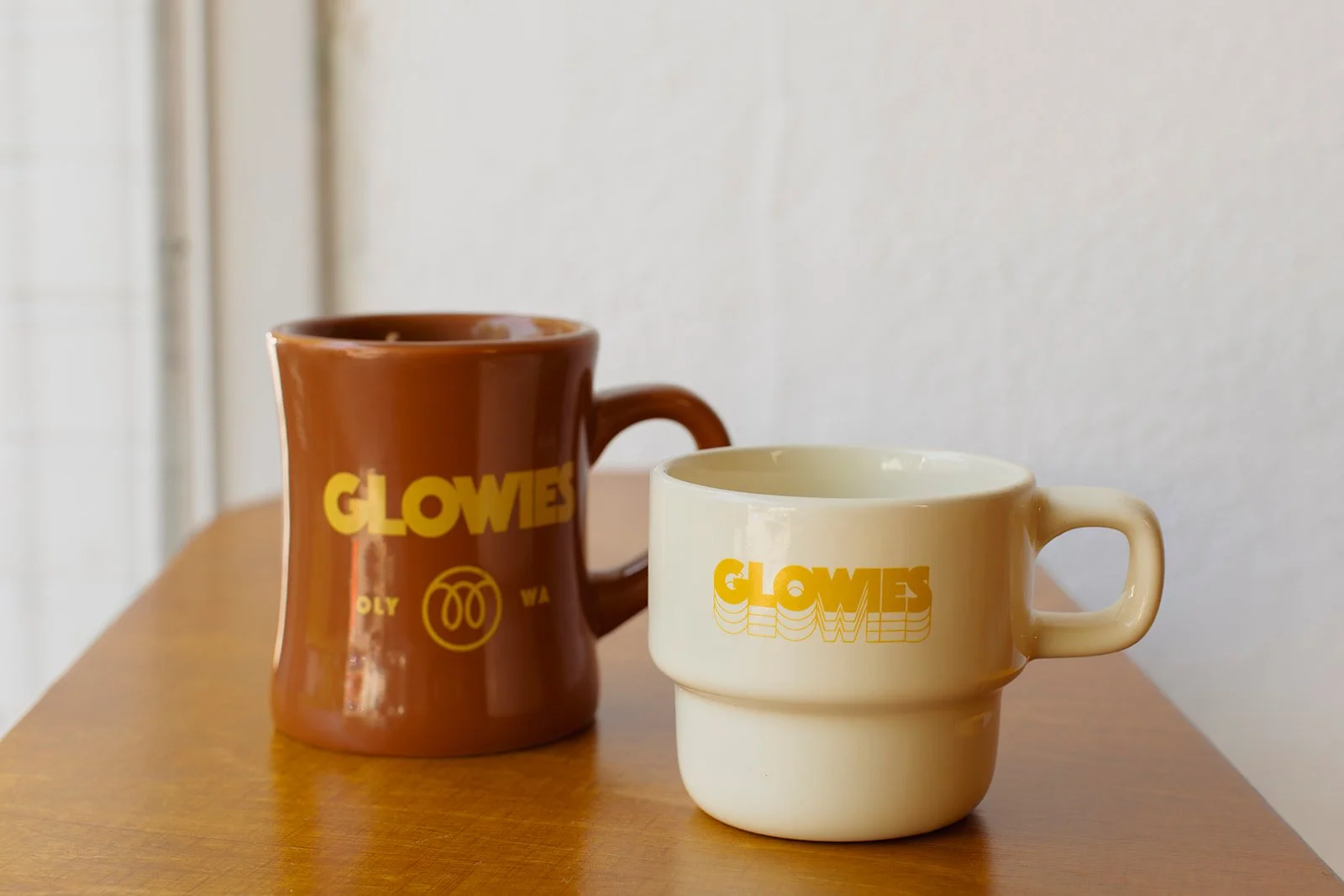 Two ceramic mugs with yellow 'GLOWES' logos, one brown and one cream, placed on a wooden surface against a plain wall.