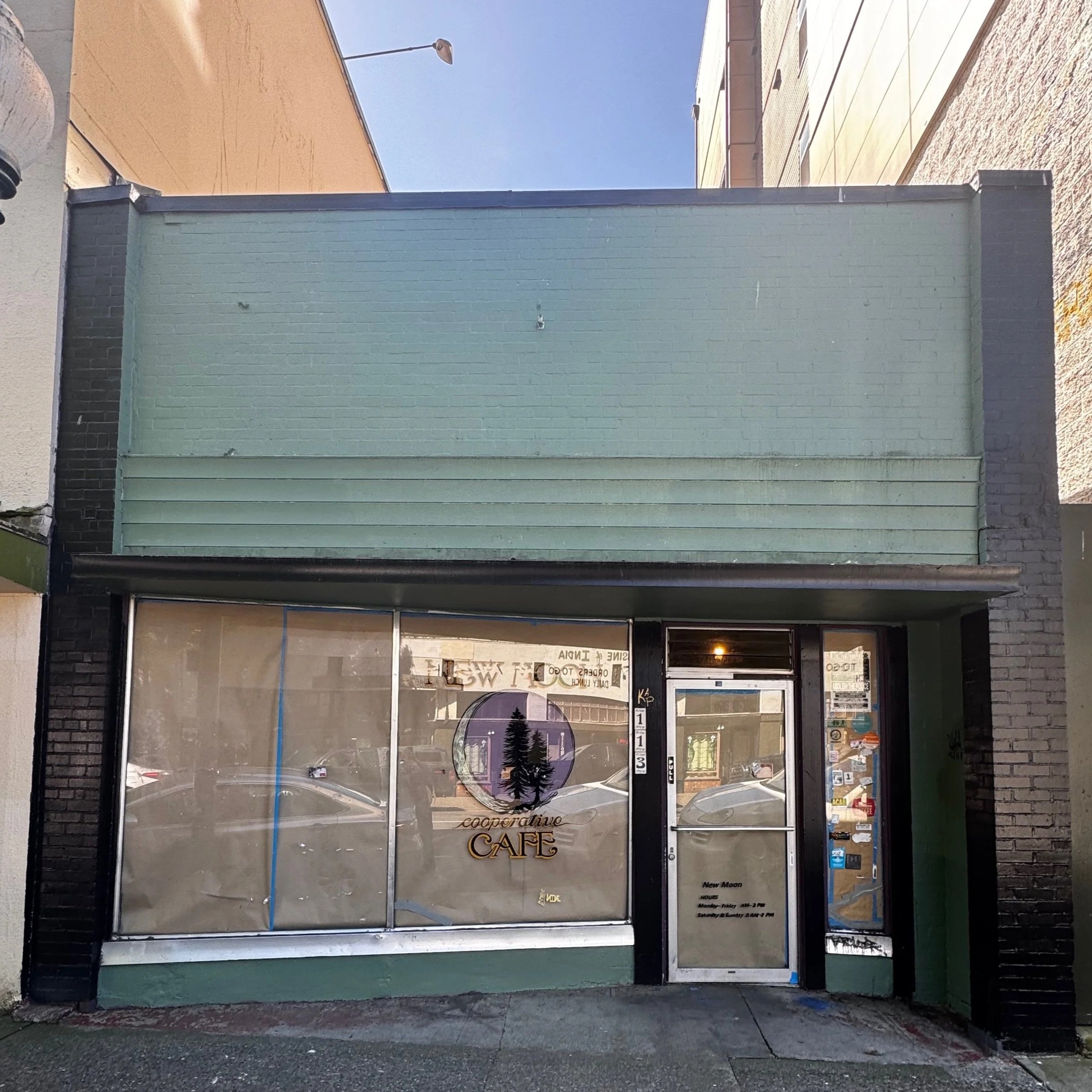 A storefront with a glass door and large window, displaying a sign that reads 'cooperative CAFE'. The door has a small round window with a pine tree silhouette. The building appears to be part of a commercial area, with visible sidewalk and neighboring structures.