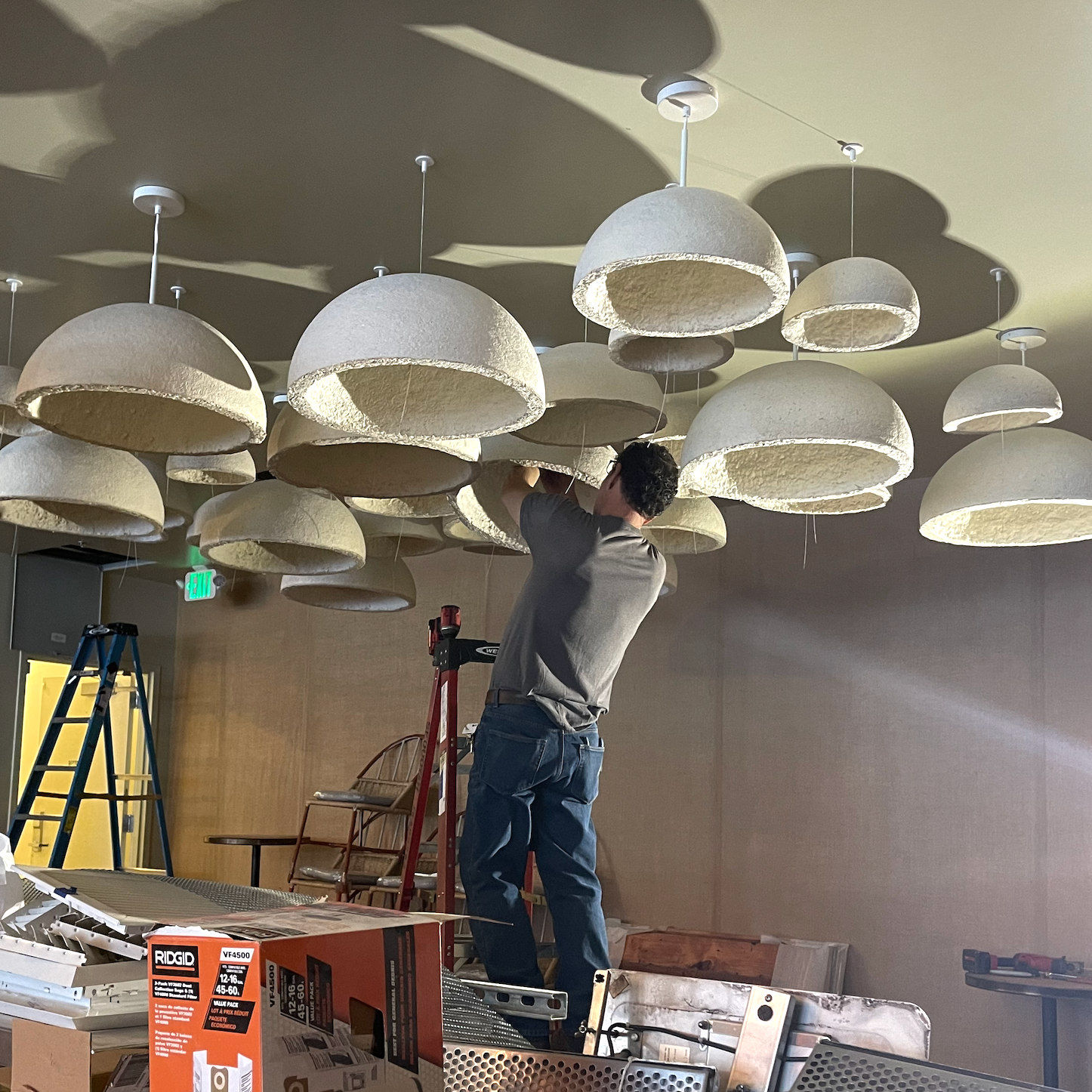 A person on a ladder installing or adjusting ceiling lights that are dome-shaped and appear to be made of a textured material, in a room with wooden walls and various tools and equipment nearby.