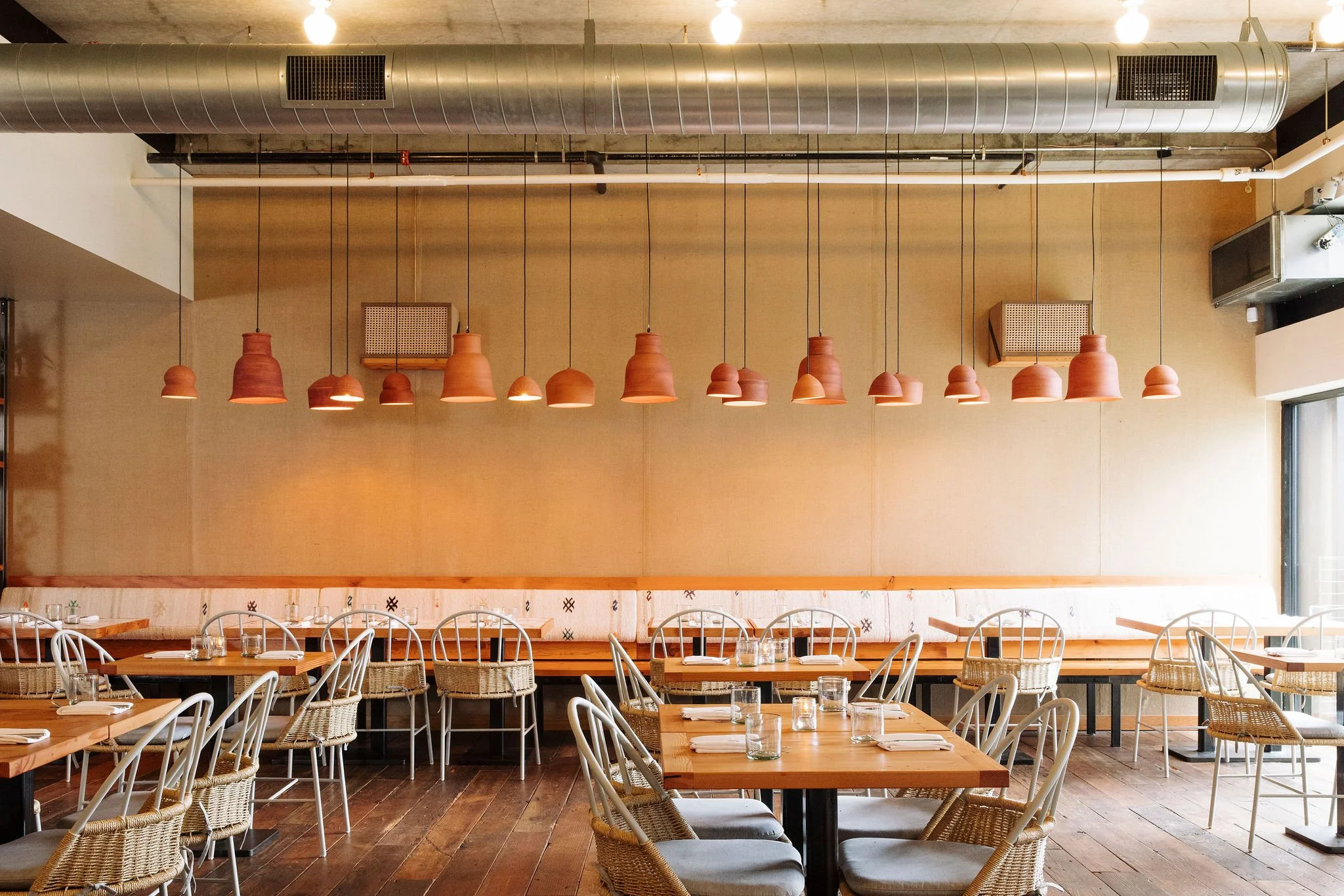 Interior of a modern restaurant with wooden tables, white chairs, and woven rattan chairs. Hanging from the ceiling are multiple red ceramic pendant lights. Air conditioning vents and exposed ductwork are visible on the ceiling. The atmosphere is bri